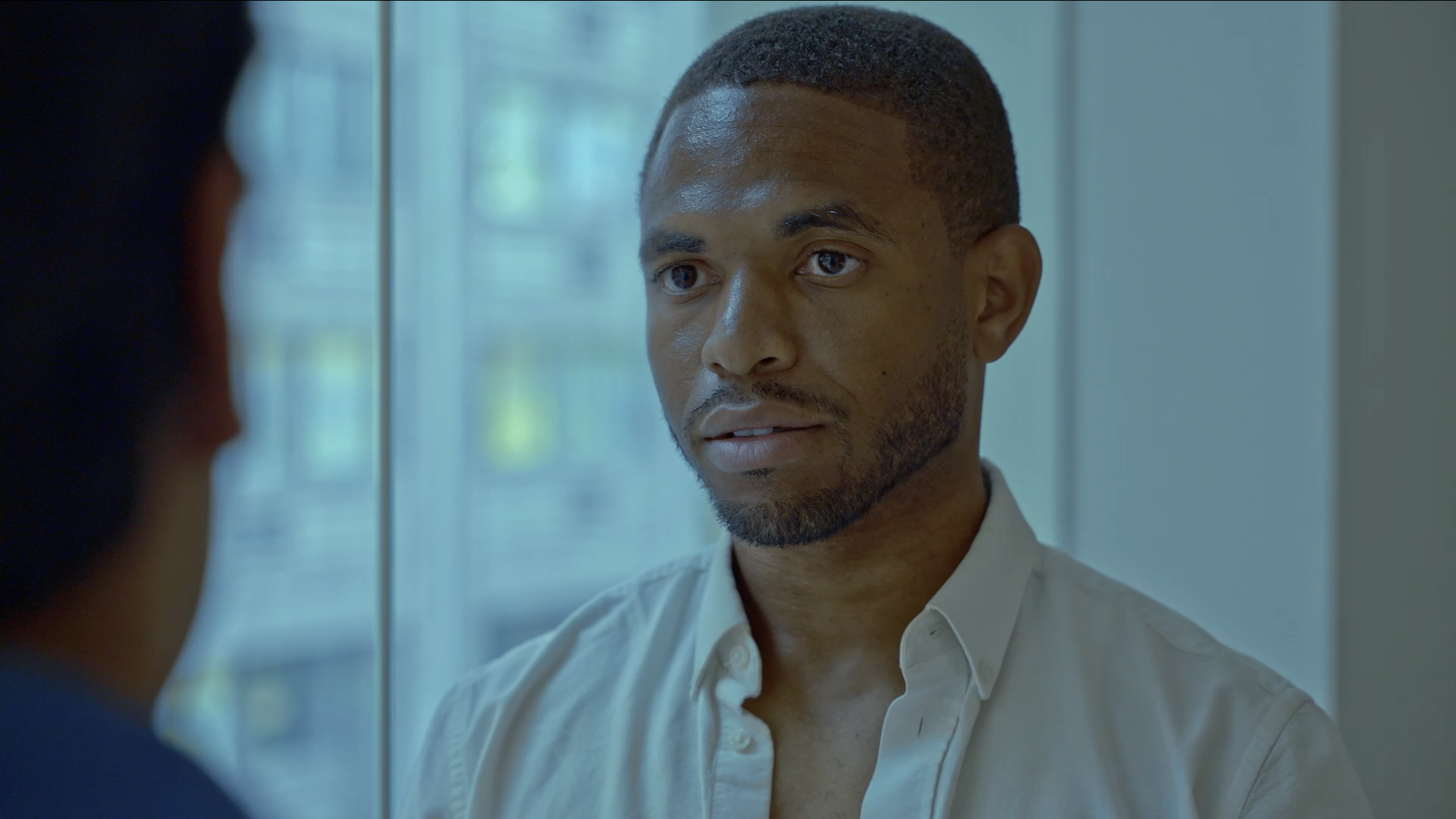 A man with short hair and a beard looking at another person through a glass window or mirror, with soft natural lighting in a bright indoor setting.