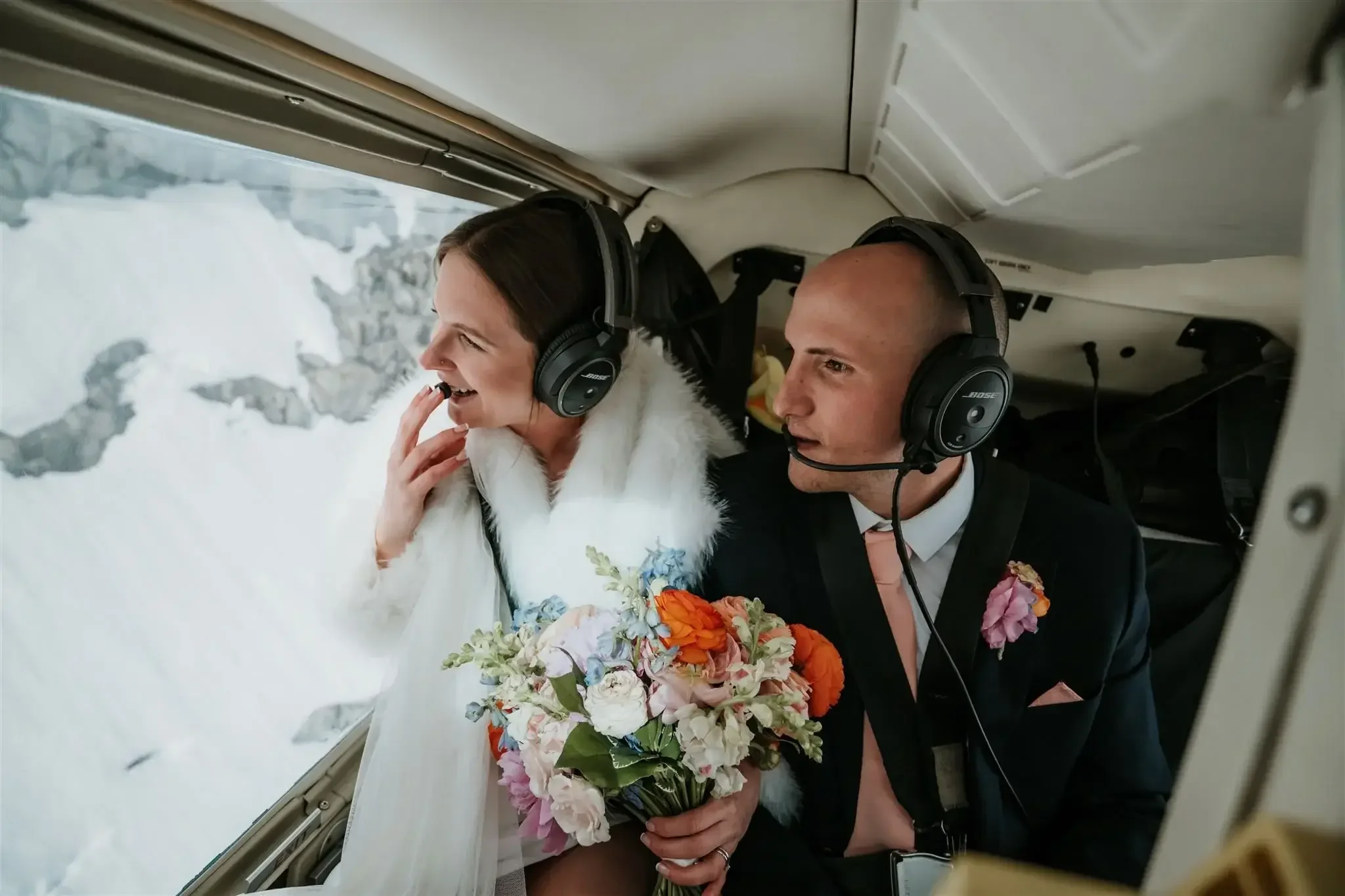 A couple on a helicopter ride, with the woman holding a bouquet, smiling and looking out the window at snow-covered mountains.