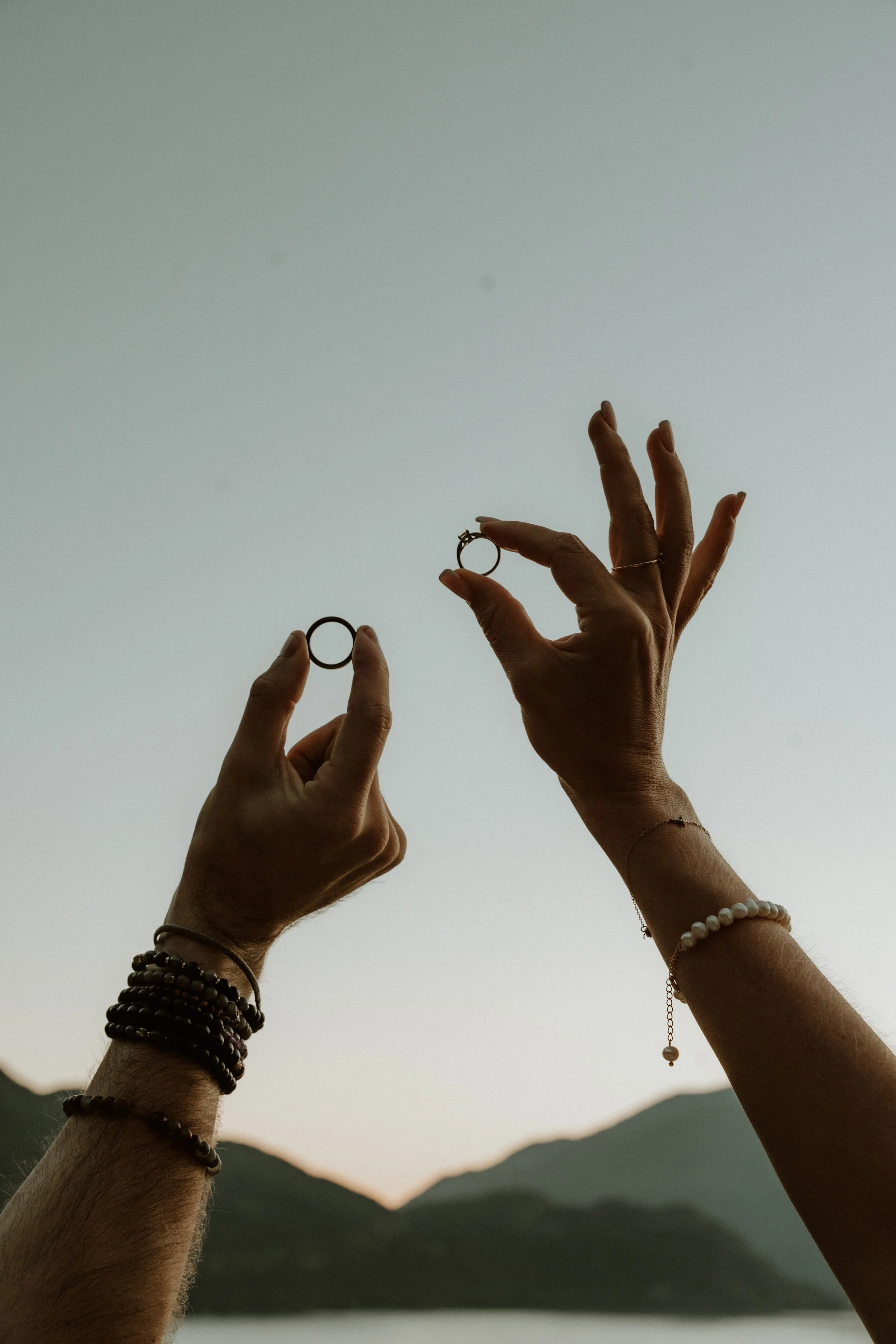 Hands holding rings against a lake and mountain backdrop during sunset or sunrise.