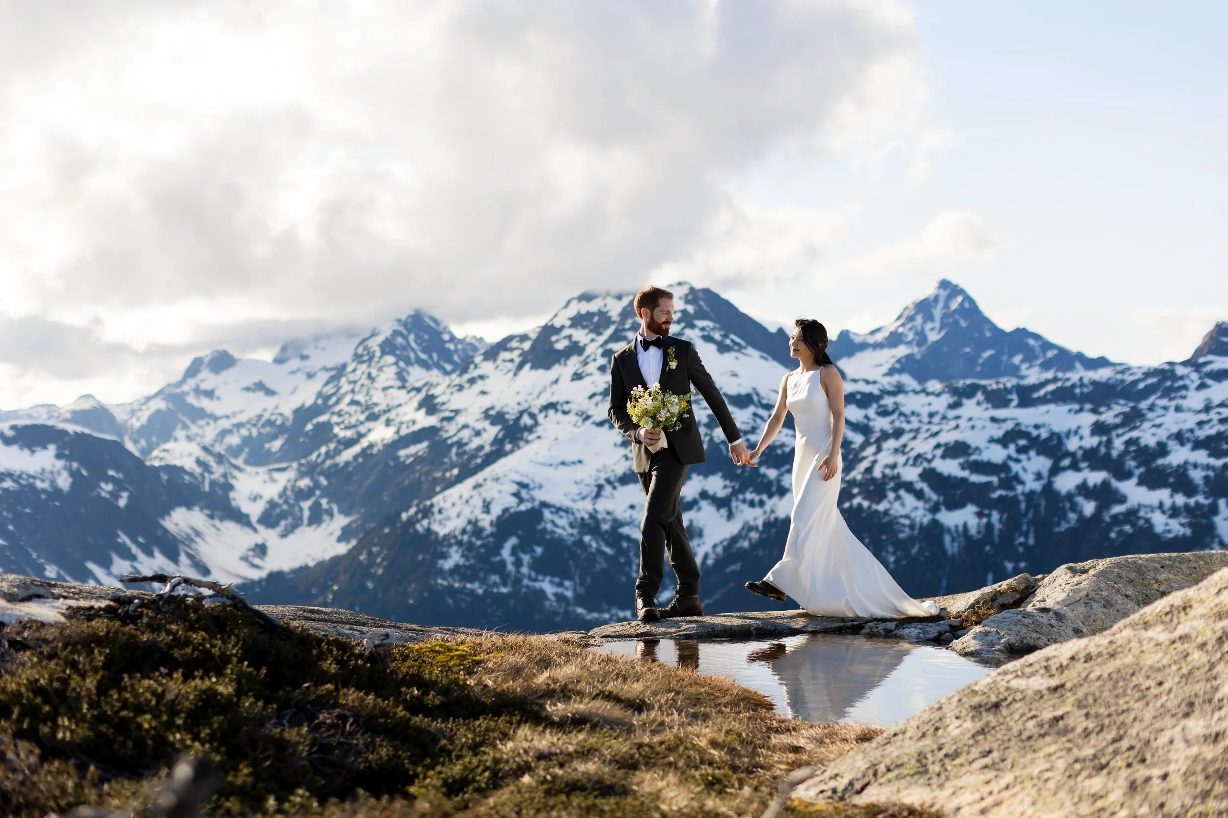 A bride and groom on a mountaintop ridge, looking at each other with snow-capped mountains in the background