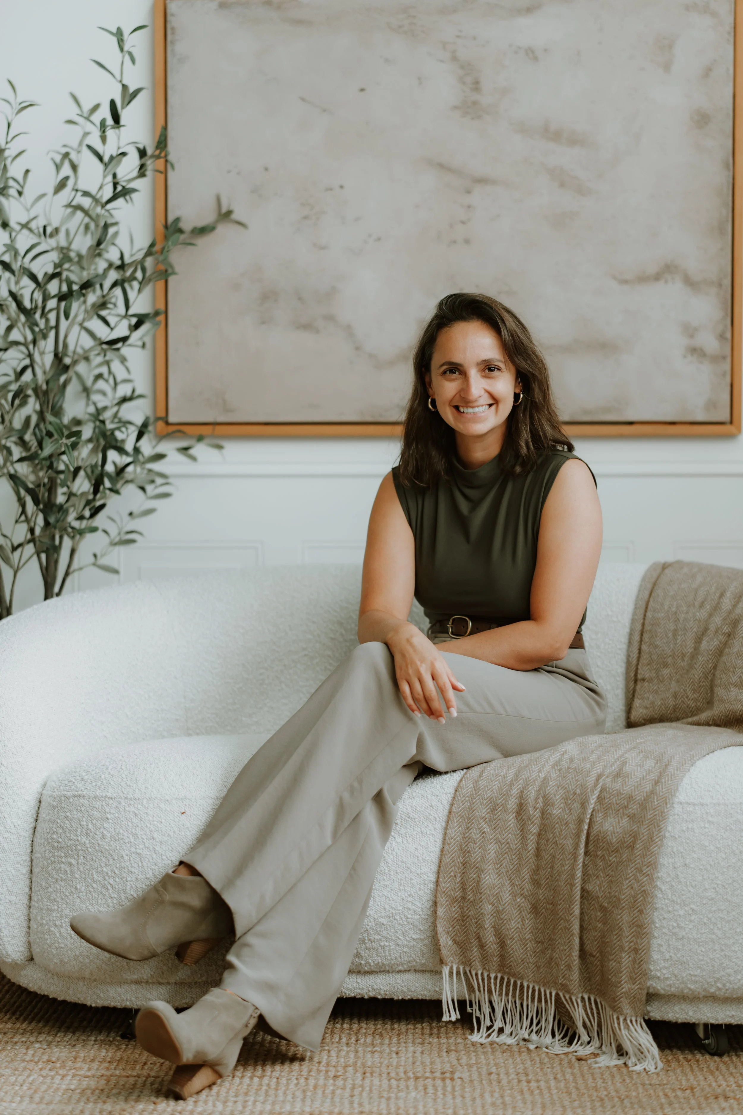 Jude, smiling, sitting on a cream-colored sofa in a room with a large abstract painting and a potted plant nearby. She wears a sleeveless green top, beige pants, and matching beige booties, with a beige blanket draped over the sofa.