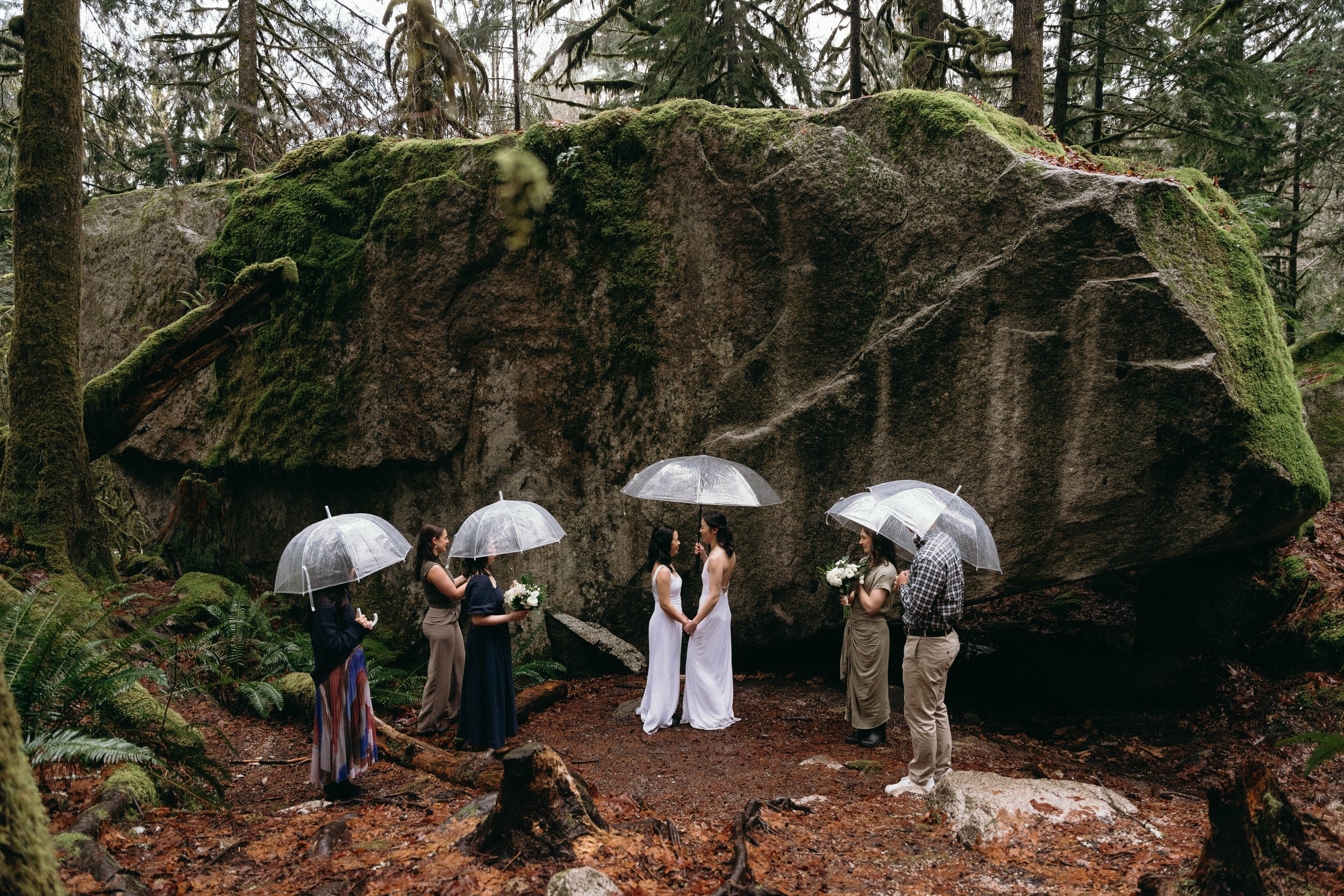 Two brides holding hands during their wedding ceremony, standing infront of a lichen covered boulder in the forest