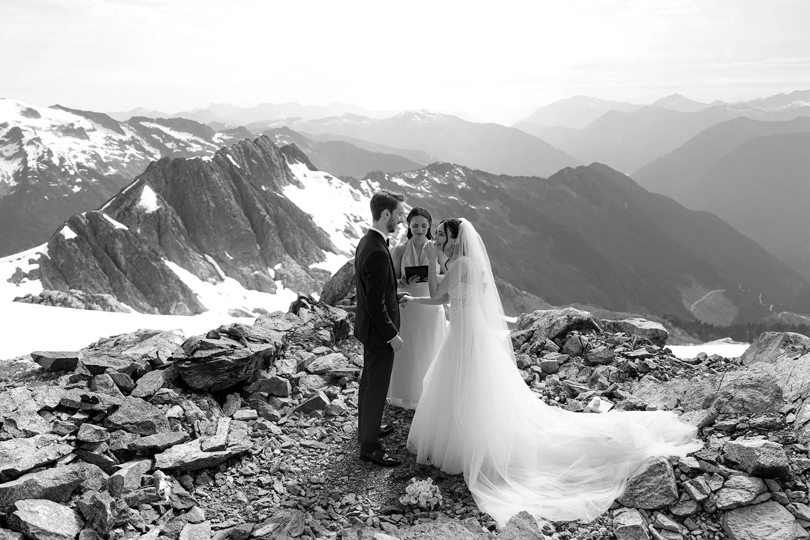 Black and white photo of a helicopter elopement in BC, with mountainous rocky terrain with snow-capped peaks in the background. A bride in a gown is holding hands with a groom in a suit as they stand facing an officiant, who is reading from a book.