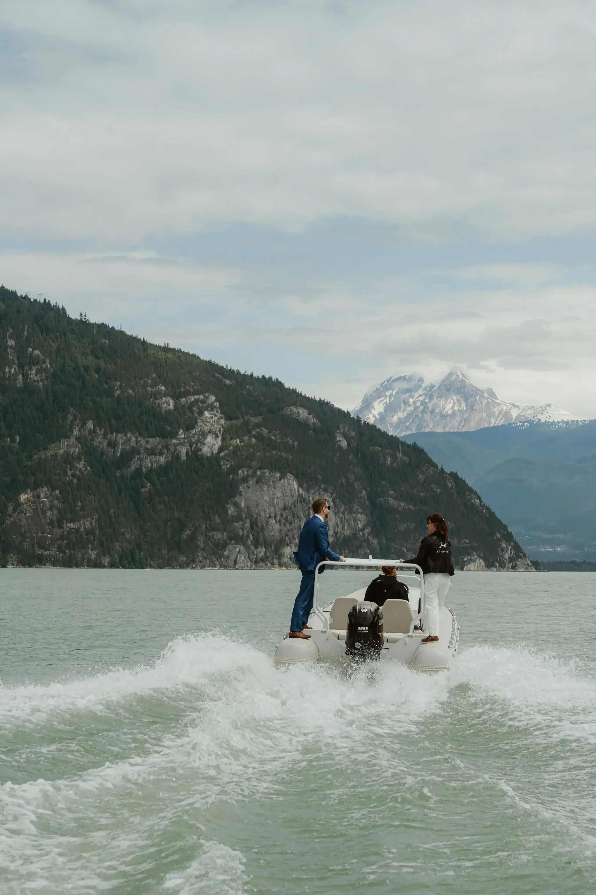 Three people on a boat in a lake surrounded by mountains and snow-capped peaks, with cloudy sky overhead.