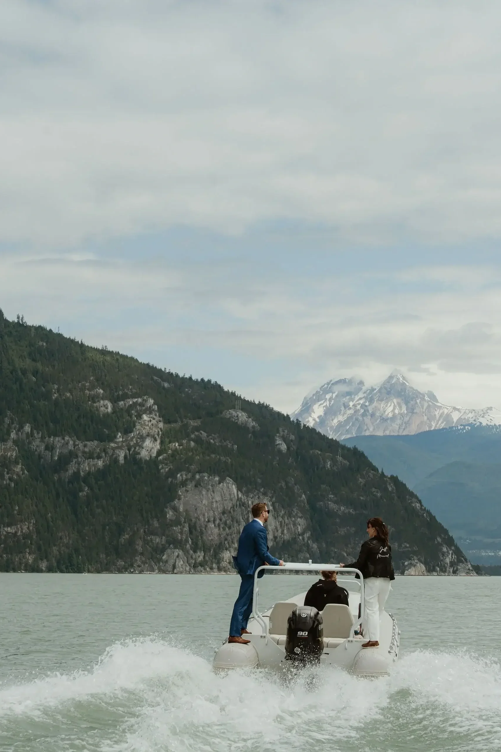 Three people on a small boat in a lake surrounded by mountains, with snow-capped peaks in the background.