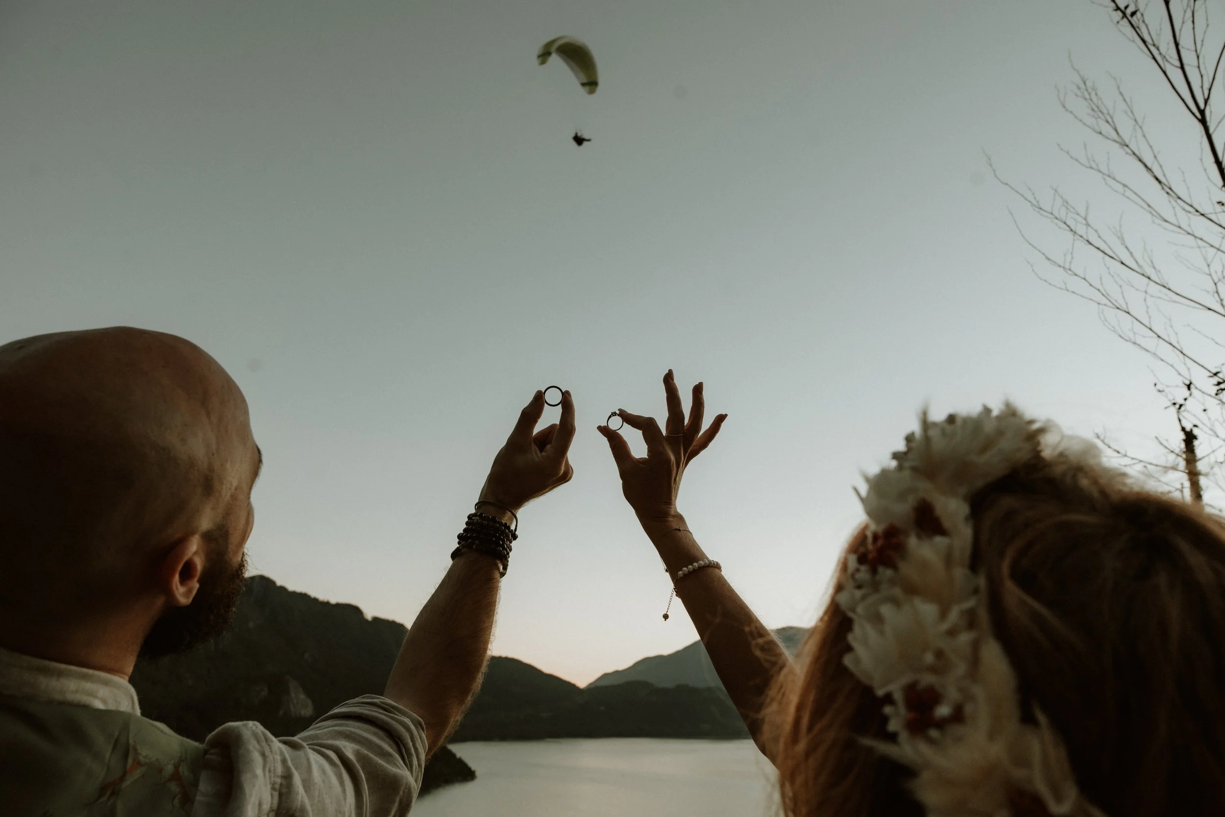 a bride and groom holding their wedding rings up at dusk, looking out over the water and mountains with a paraglider in the background.