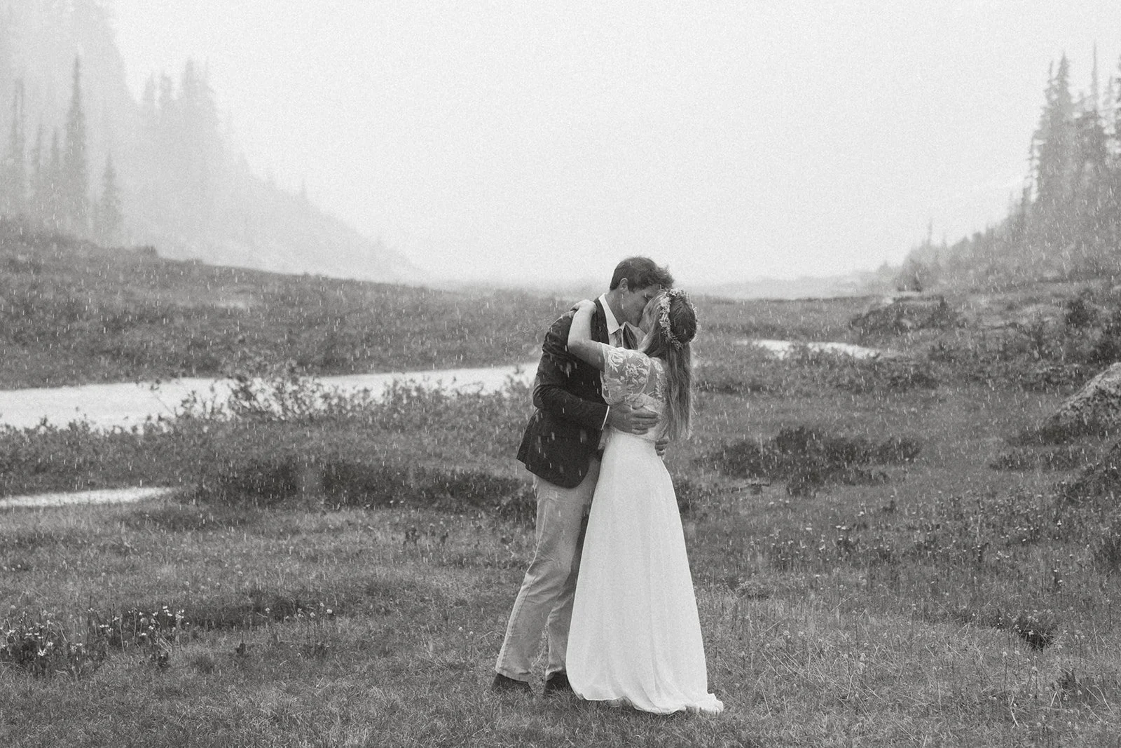 A bride and groom sharing a kiss in the rain on a grassy field with a mountain landscape in the background.