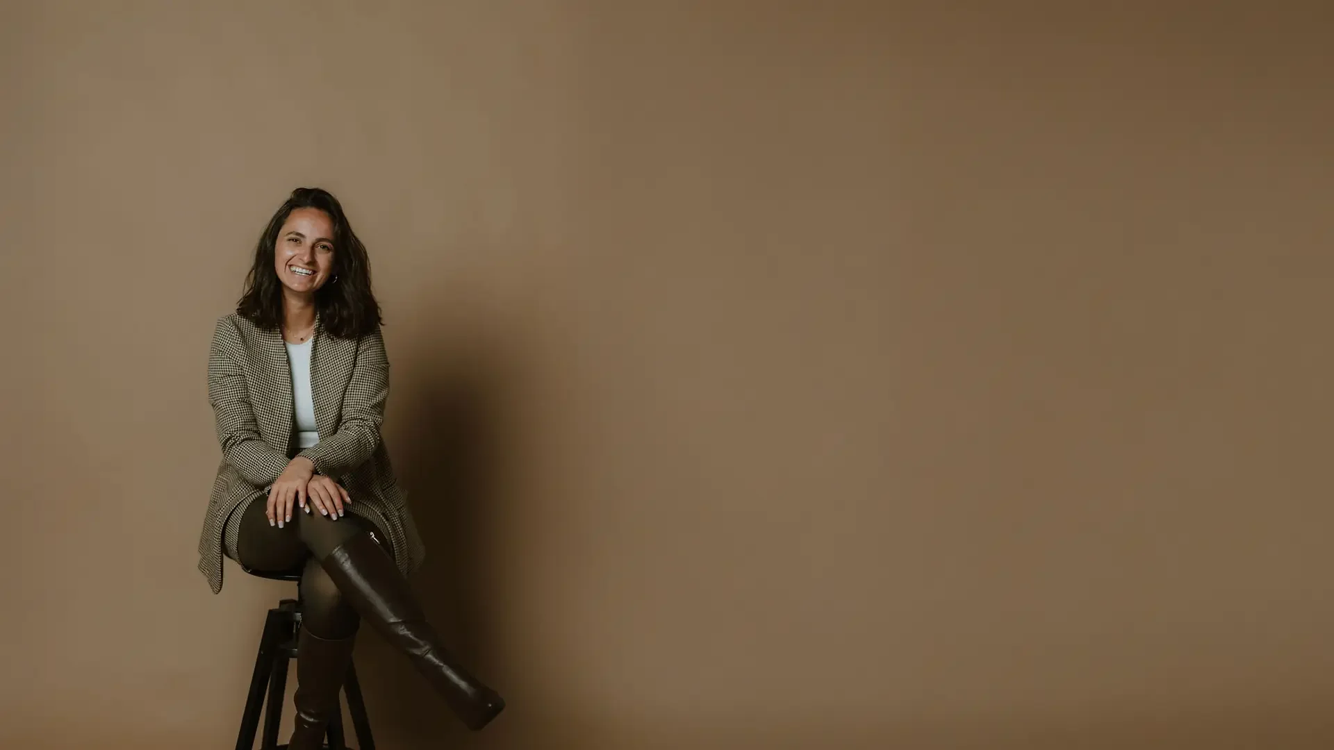 A woman with shoulder-length brown hair, smiling, sitting on a black stool against a plain brown background. She is wearing a brown checkered blazer, a white top, black leggings, and brown knee-high boots.