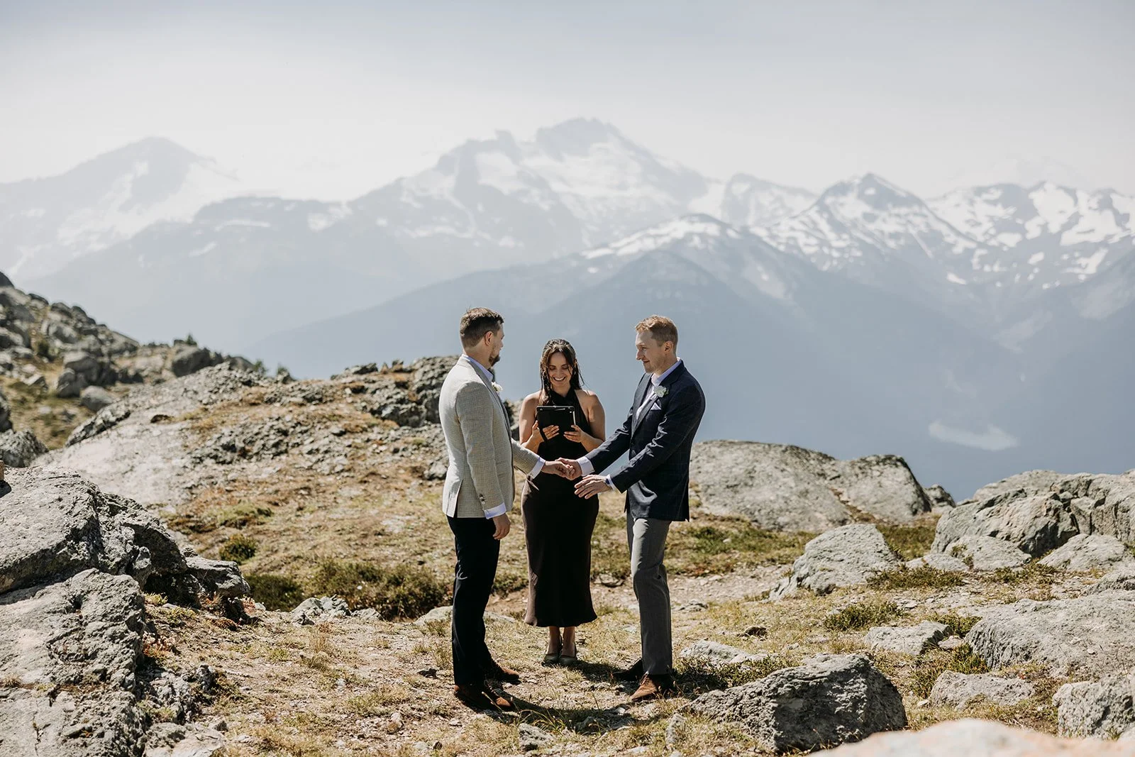 Two grooms stand on top of a mountain in their wedding ceremony with their officiant between them