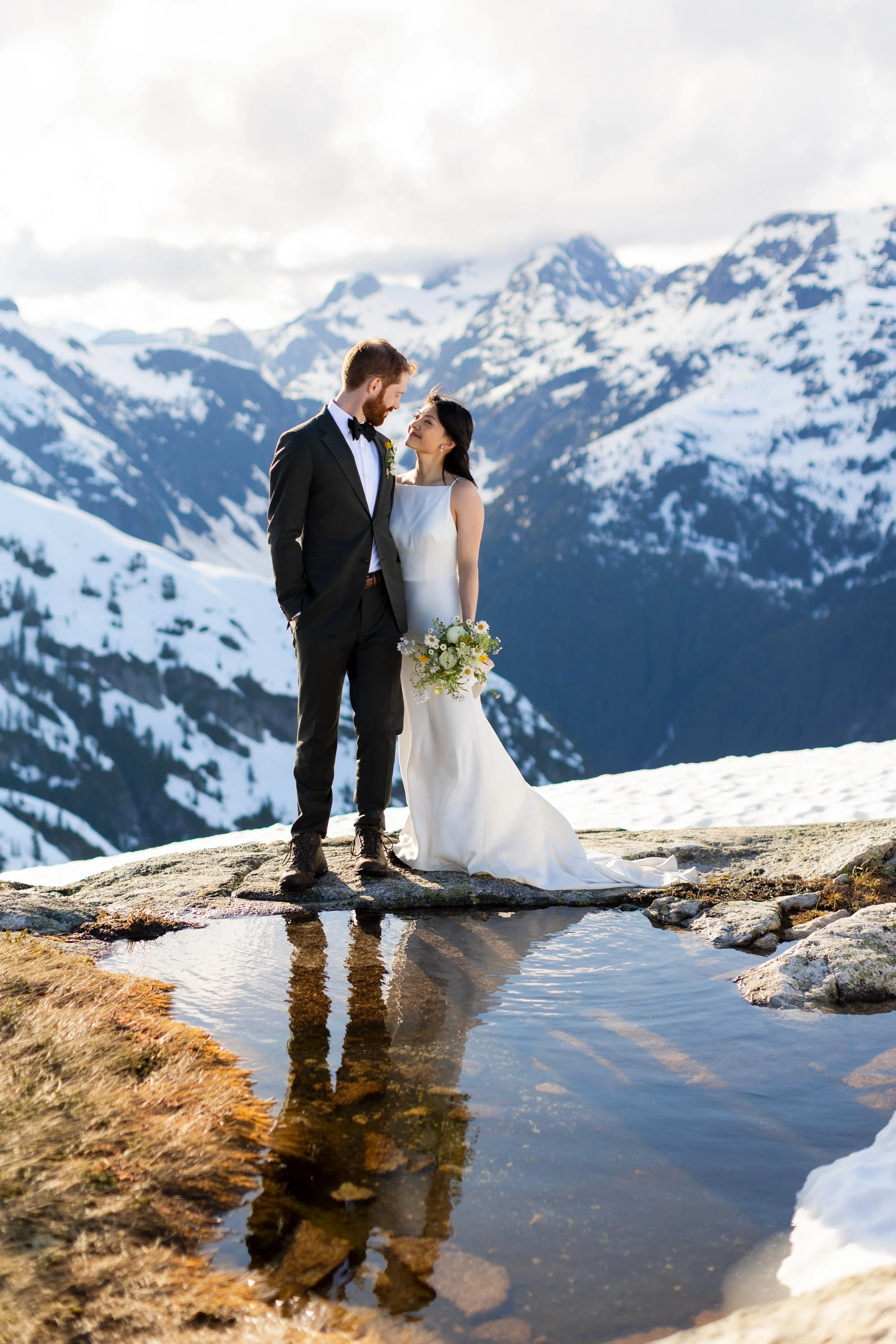 A bride and groom standing on top of a mountain on their wedding day, their reflection is in a puddle and the mountains are behind them