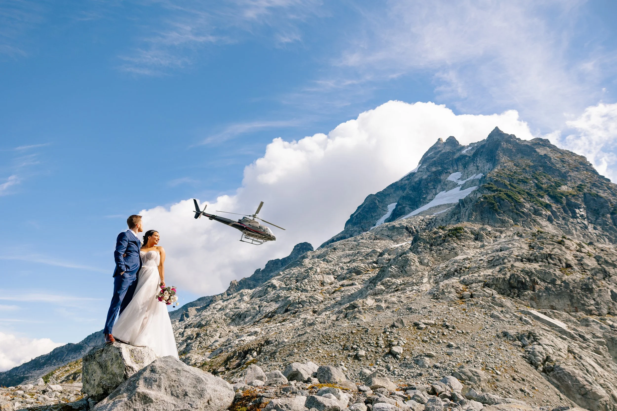 A couple in their wedding clothes with a rugged peak and helicopter in the background