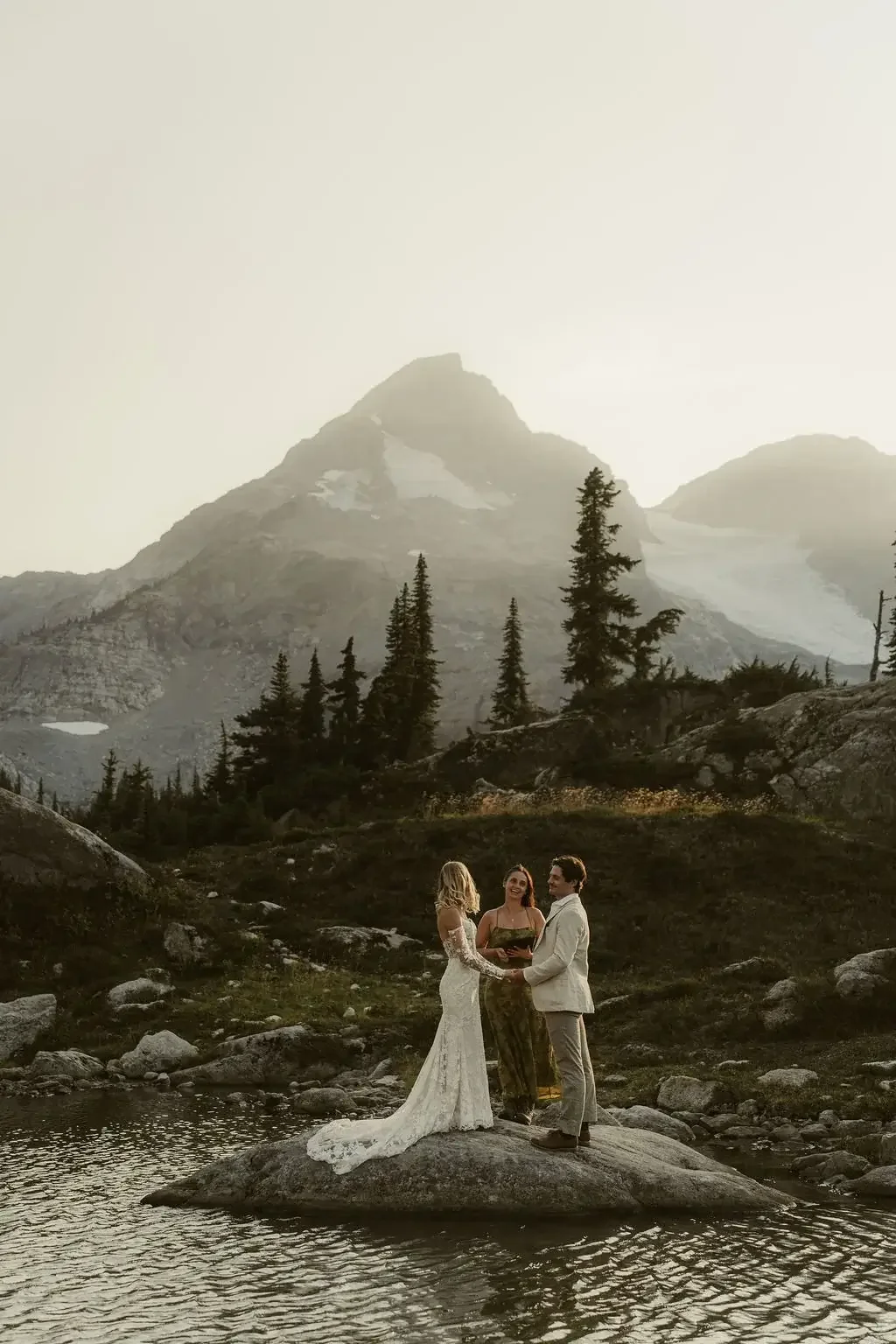 A wedding ceremony taking place outdoors on a rock by a river, with a mountain and trees in the background, during sunset or sunrise.