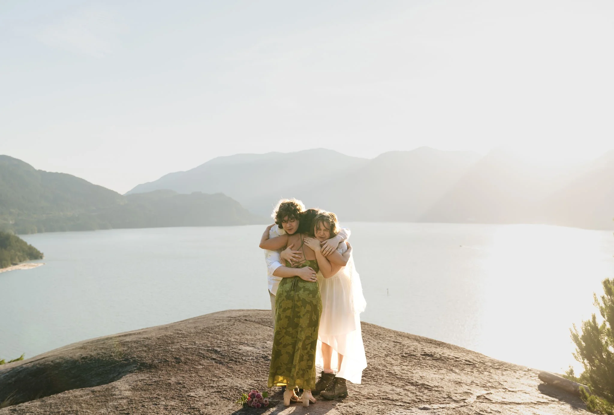 Jude and couple hugging on a rocky ledge overlooking a body of water with mountains in the background, with the sun setting or rising.