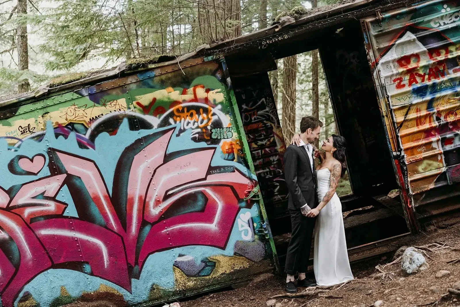 A bride and groom, holding hands and gazing at each other, standing in front of a graffiti-covered, tilted metal container in a wooded area.