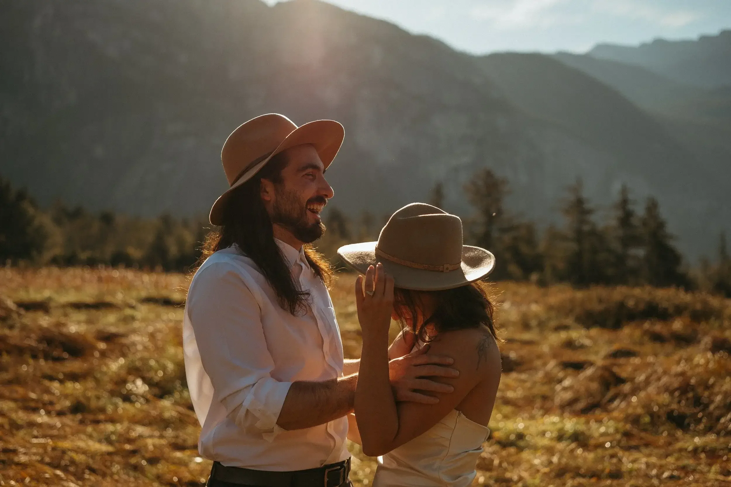 Jude and Virgile in a field outdoors, wearing wide-brimmed hats, smiling and engaging with each other, with mountains in the background at sunset.