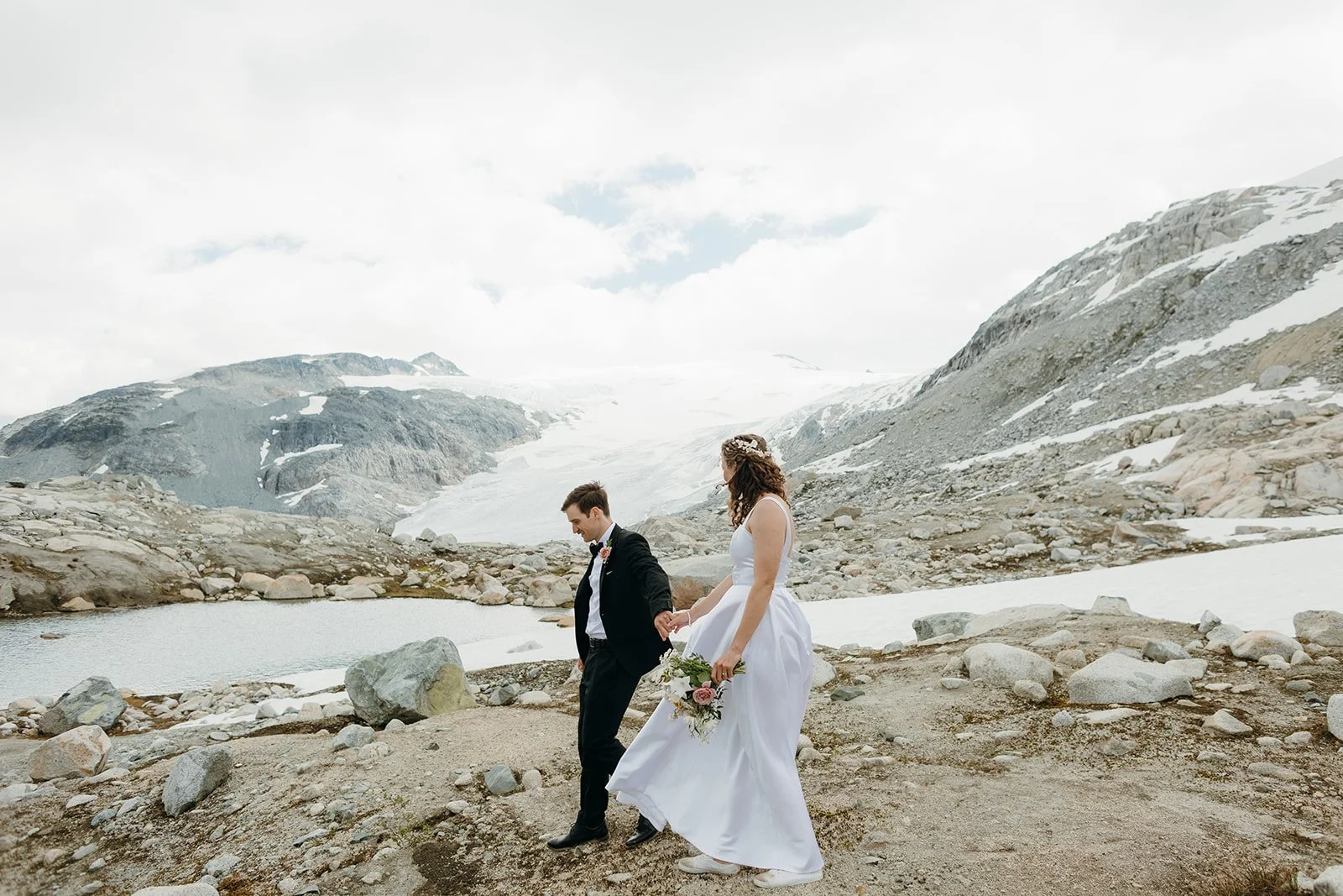 A bride and groom holding hands as they walk through the alpine, a lake and glacier behind them