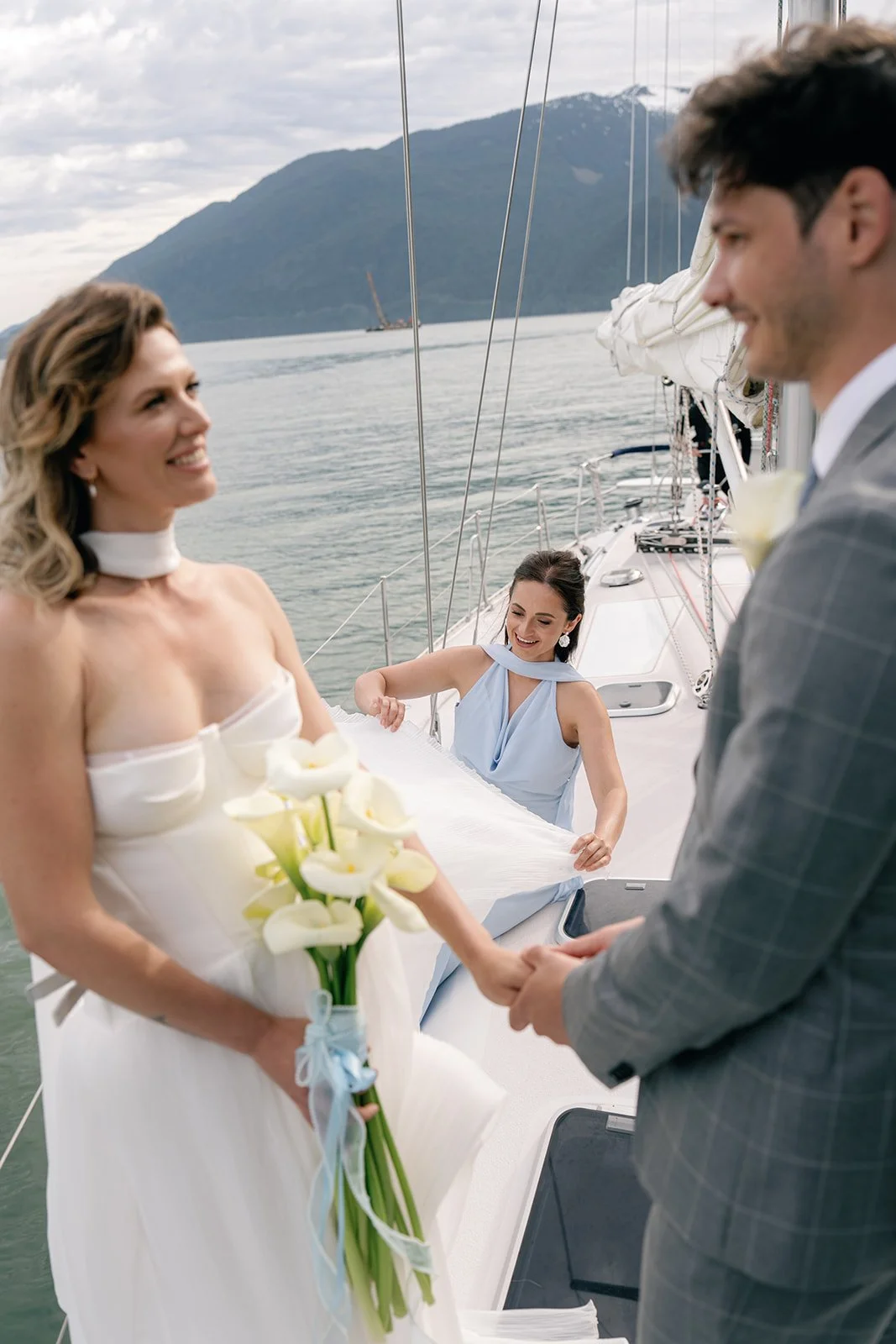 a bride and groom on a sailboat, holding hands, while their elopement planner fixes her bridal gown