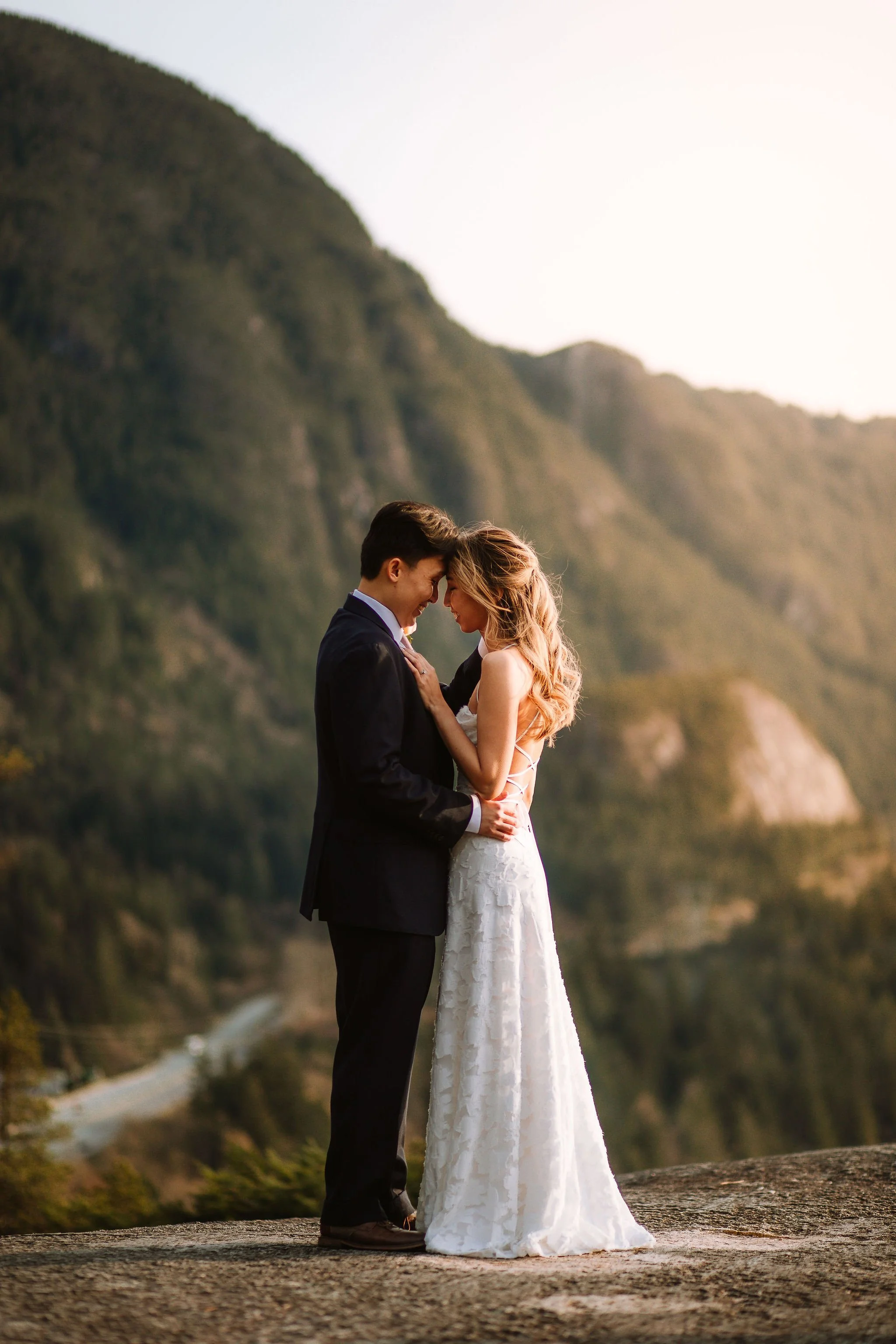 A bride and groom standing upon a mountain top, with the Canadian mountains in the background, wearing a suit and white dress, embracing