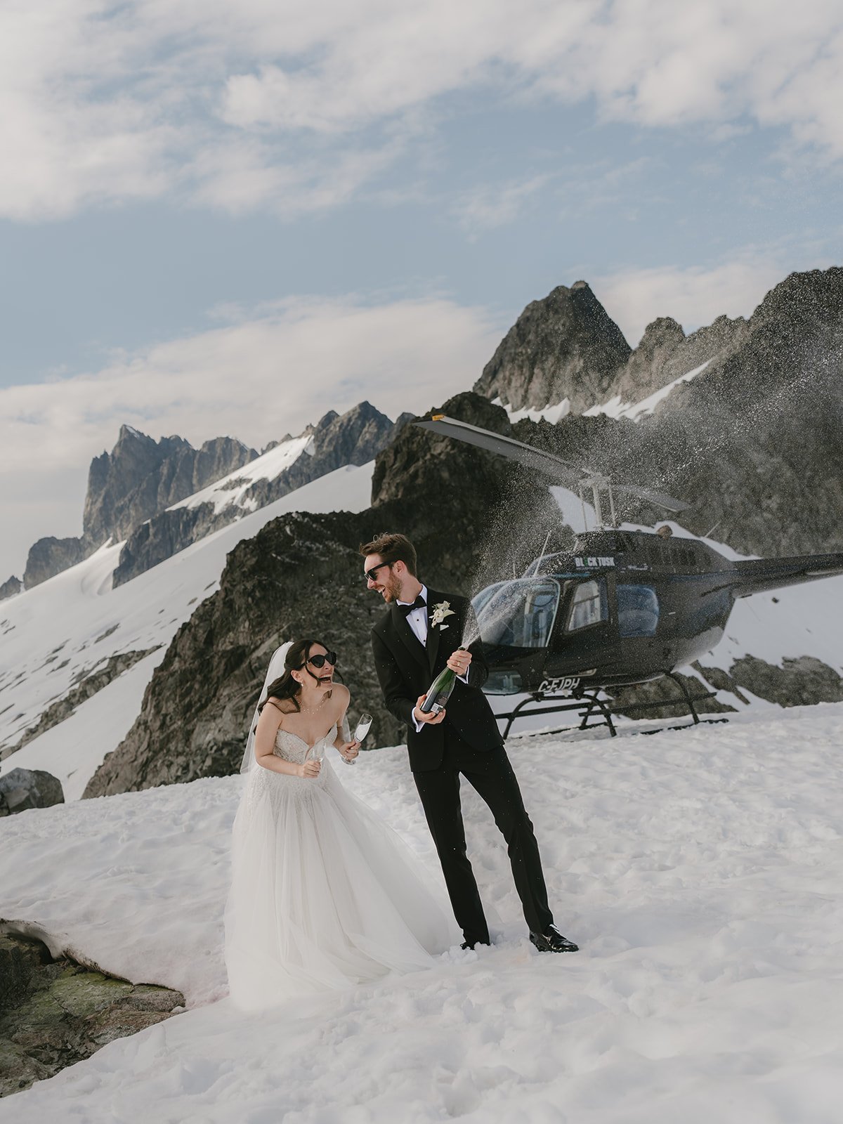 A photo from a helicopter elopement, with a bride and groom in wedding attire celebrating in a snowy mountain landscape, with a helicopter in the background and one of them holding a champagne bottle.