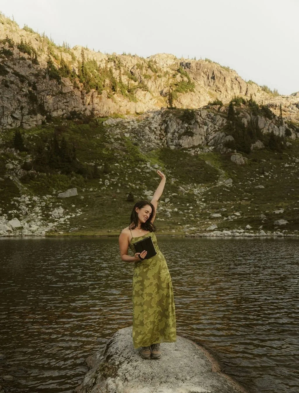 A woman in a green dress stands on a rock in a lake, holding a book and raising one arm in a scenic mountain landscape.