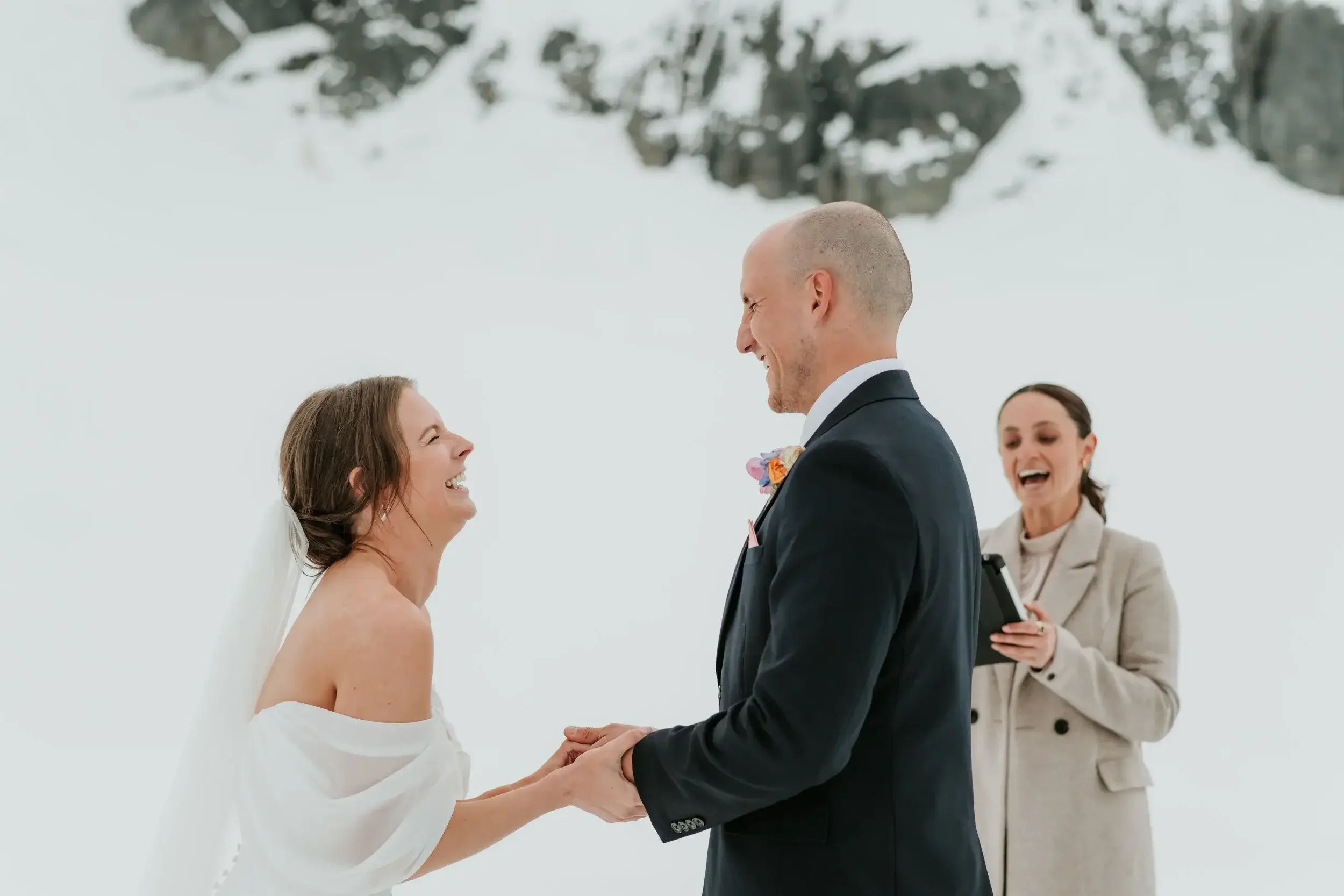A bride and groom smiling and holding hands during their wedding ceremony in a snowy outdoor setting, with jude the officiant in the background.