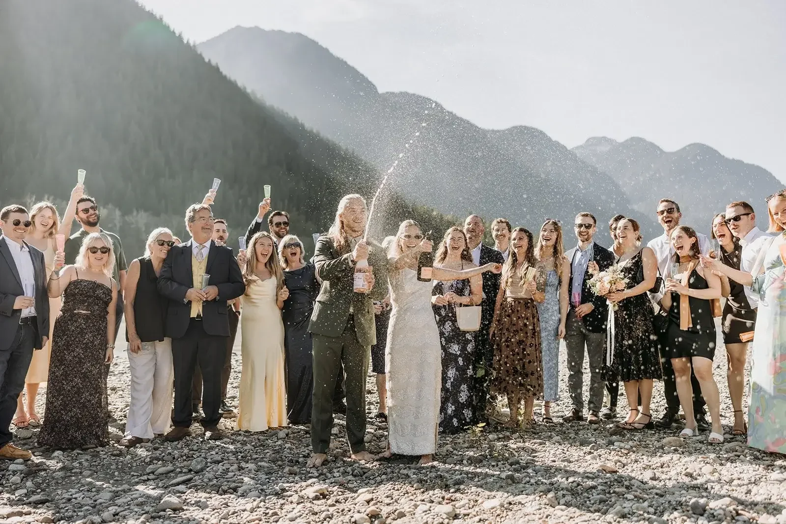 Wedding celebration on a rocky beach with a large group of people, including a bride and groom spraying champagne, with mountains in the background.