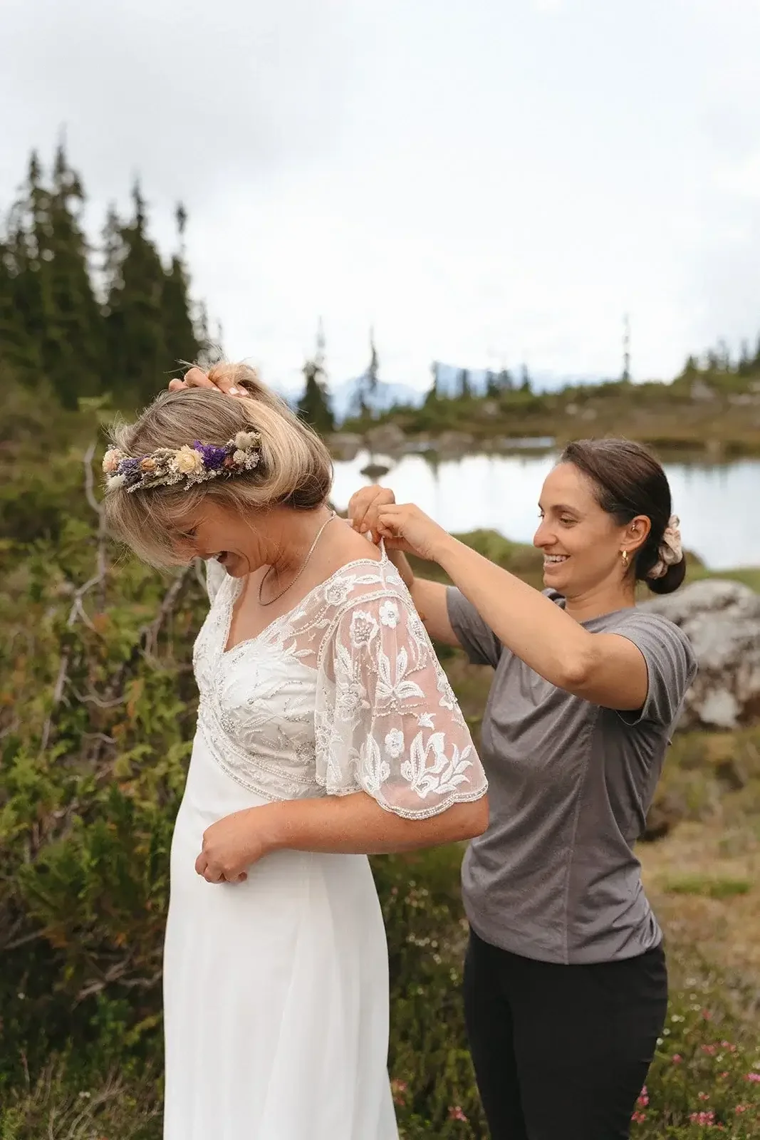 Jude the officiant in a  gray shirt helps a bride in a white dress with lace sleeves and floral crown into her dress outdoors near a lake and trees.