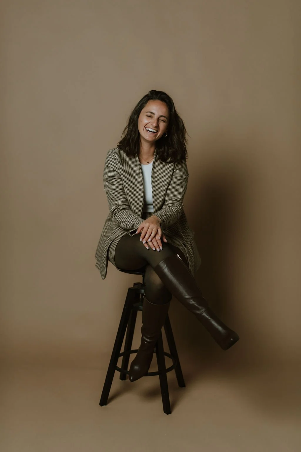 Jude, sitting on a black stool against a plain beige background, smiling and laughing. She is wearing a checked blazer, a white top, dark tights, and knee-high brown boots.