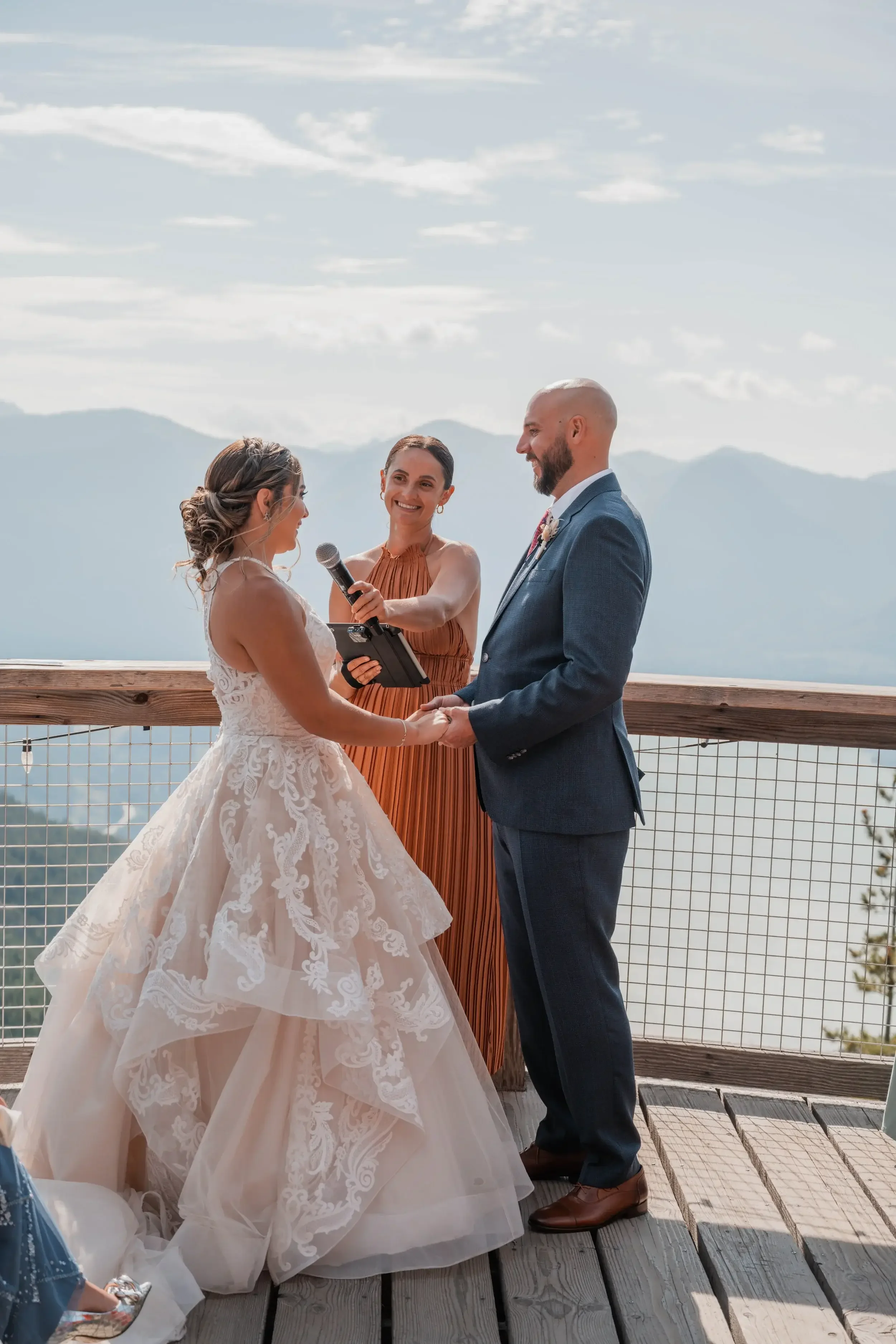A couple getting married outdoors on a wooden platform with mountain views in the background. The bride and groom are holding hands, jude the officiant is smiling, and the bride is speaking into a microphone.