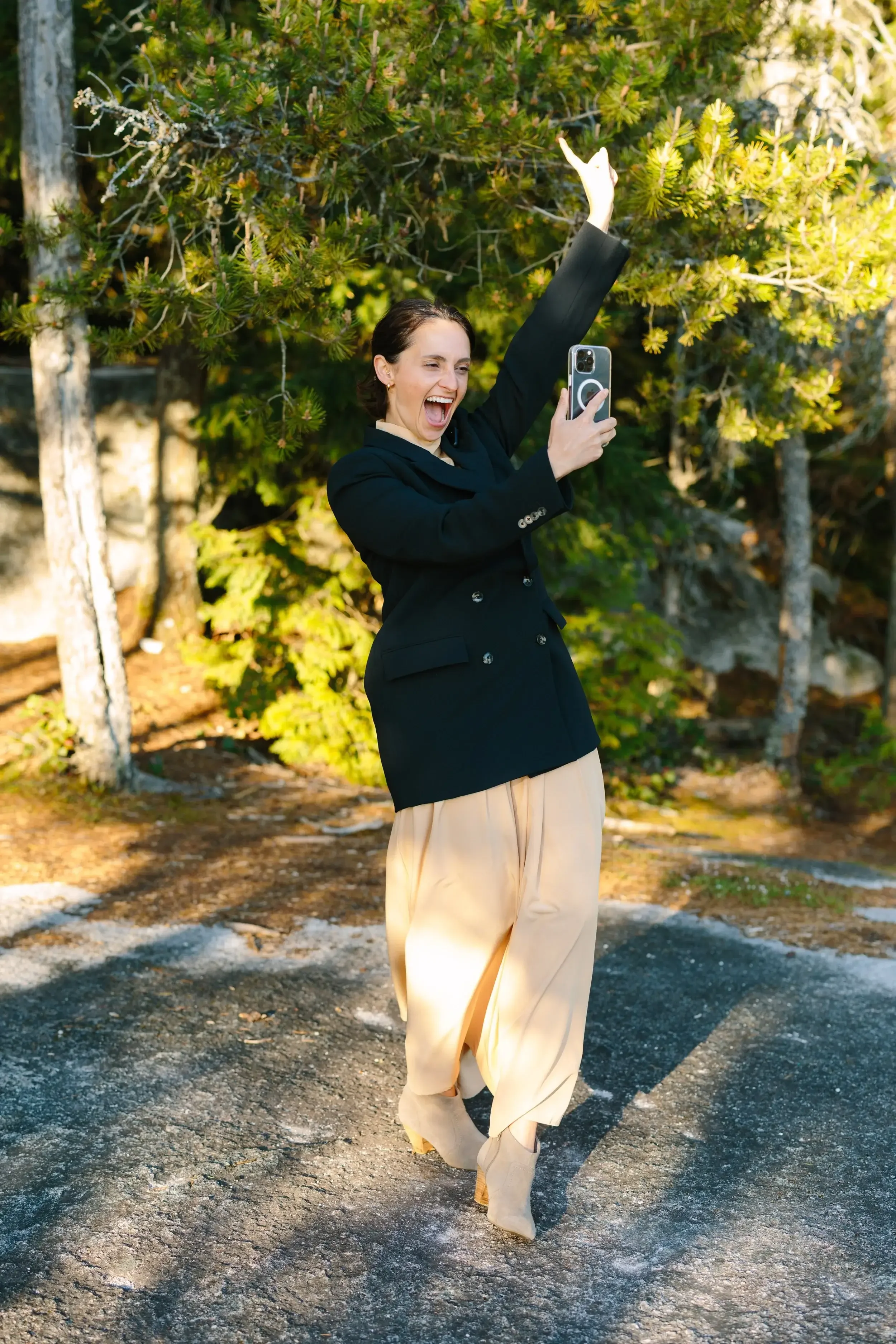 Jude outdoors on a dirt path, taking a selfie with a smartphone, surrounded by trees, smiling and appearing joyful.