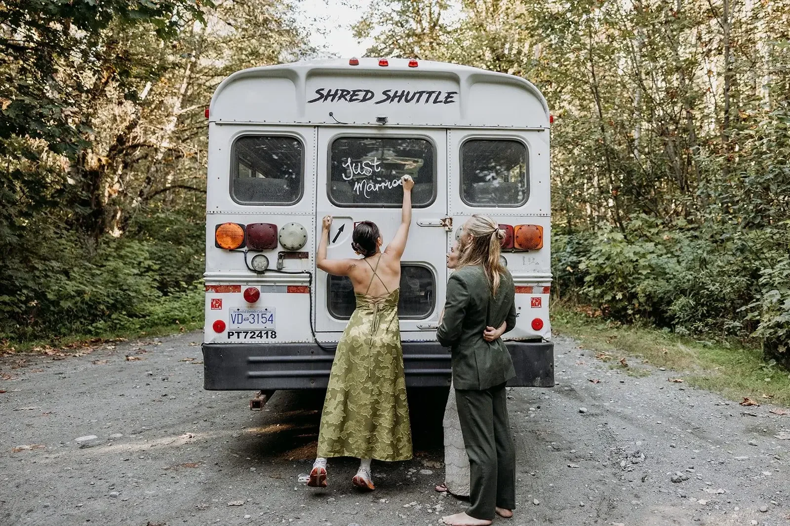 jude and bride and groom standing behind a white bus with the words 'Shred Shuttle' on it, with jude writing on the bus window and the couple watching, in a wooded outdoor area.