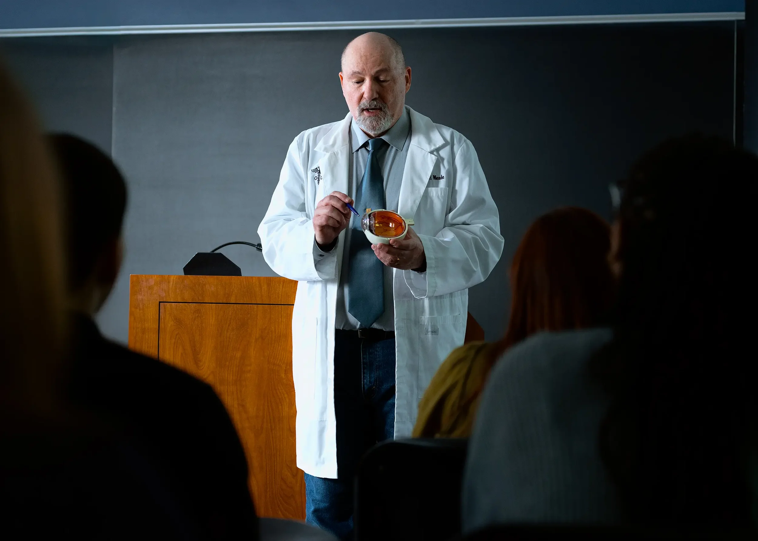 A male doctor in a white labcoat presents to a group of people in a classroom or lecture hall, holding a model of the human eye | Dr. Robert Meade | Public Speaker & Vision Consultant | Presentations about Eyes and Vision