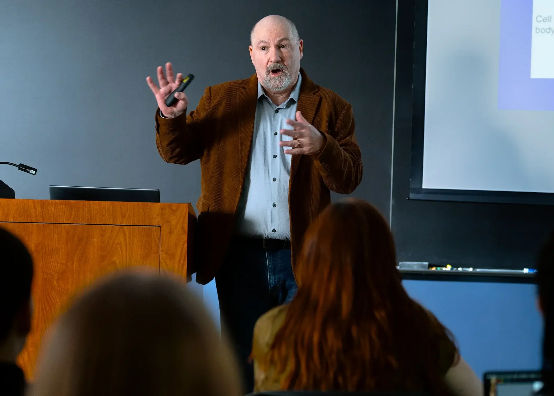 A man with a gray beard and mustache wearing a light blue shirt and brown blazer is giving a presentation in a classroom in front of a projector screen | Dr. Robert Meade | Public Speaker & Vision Consultant | Presentations about Eyes and Vision