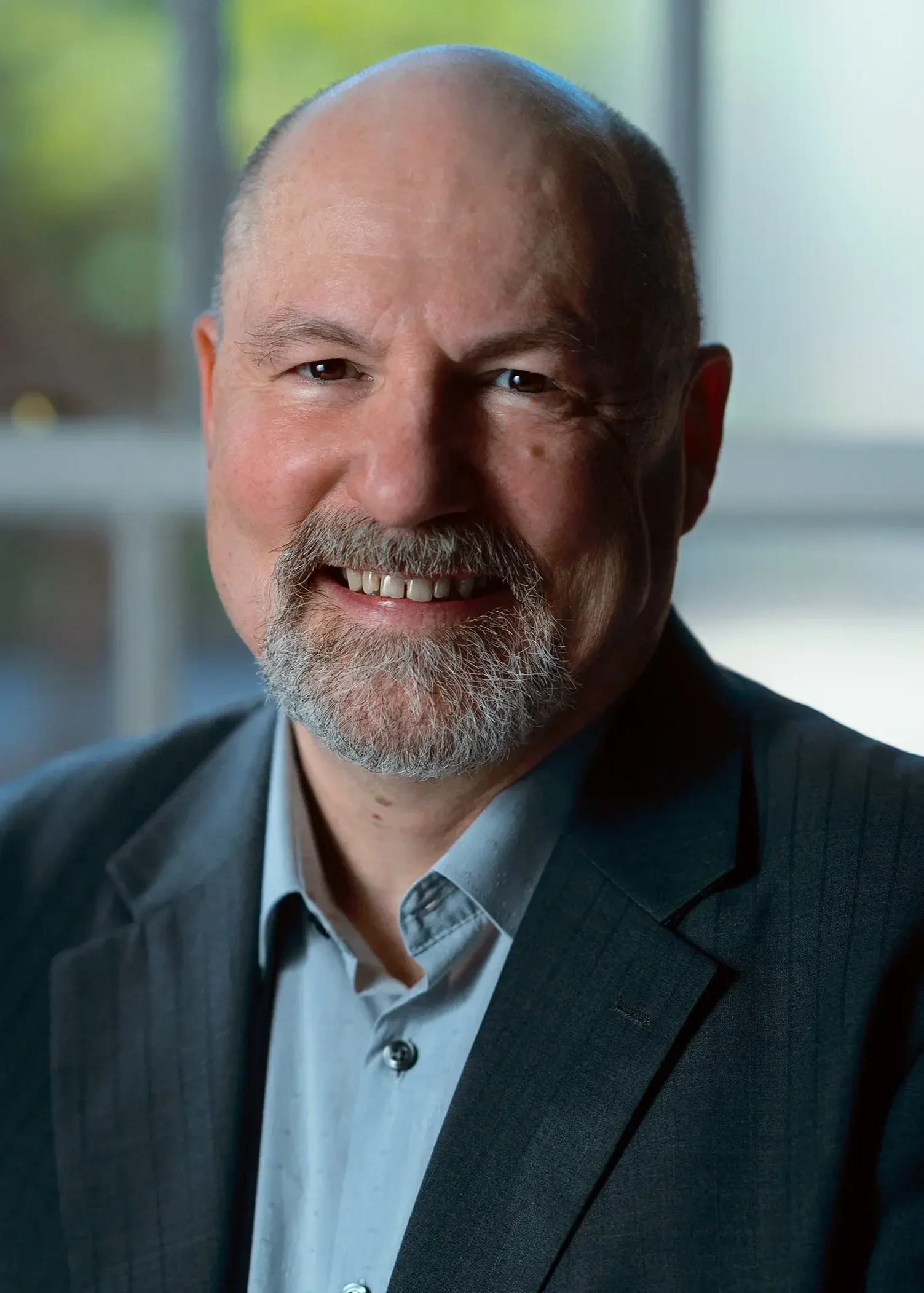 Close-up portrait of a smiling middle-aged man with a bald head, gray beard, wearing a dark suit and light blue shirt, indoors with a blurred background | Dr. Robert Meade | Public Speaker & Vision Consultant | Presentations about Eyes and Vision