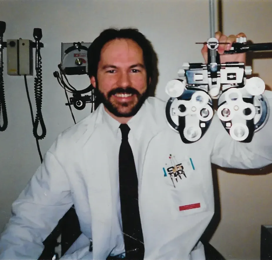 A smiling eye doctor or optometrist in a white labcoat holding a phoropter in a medical examination room | Dr. Robert Meade | Public Speaker & Vision Consultant | Presentations about Eyes and Vision