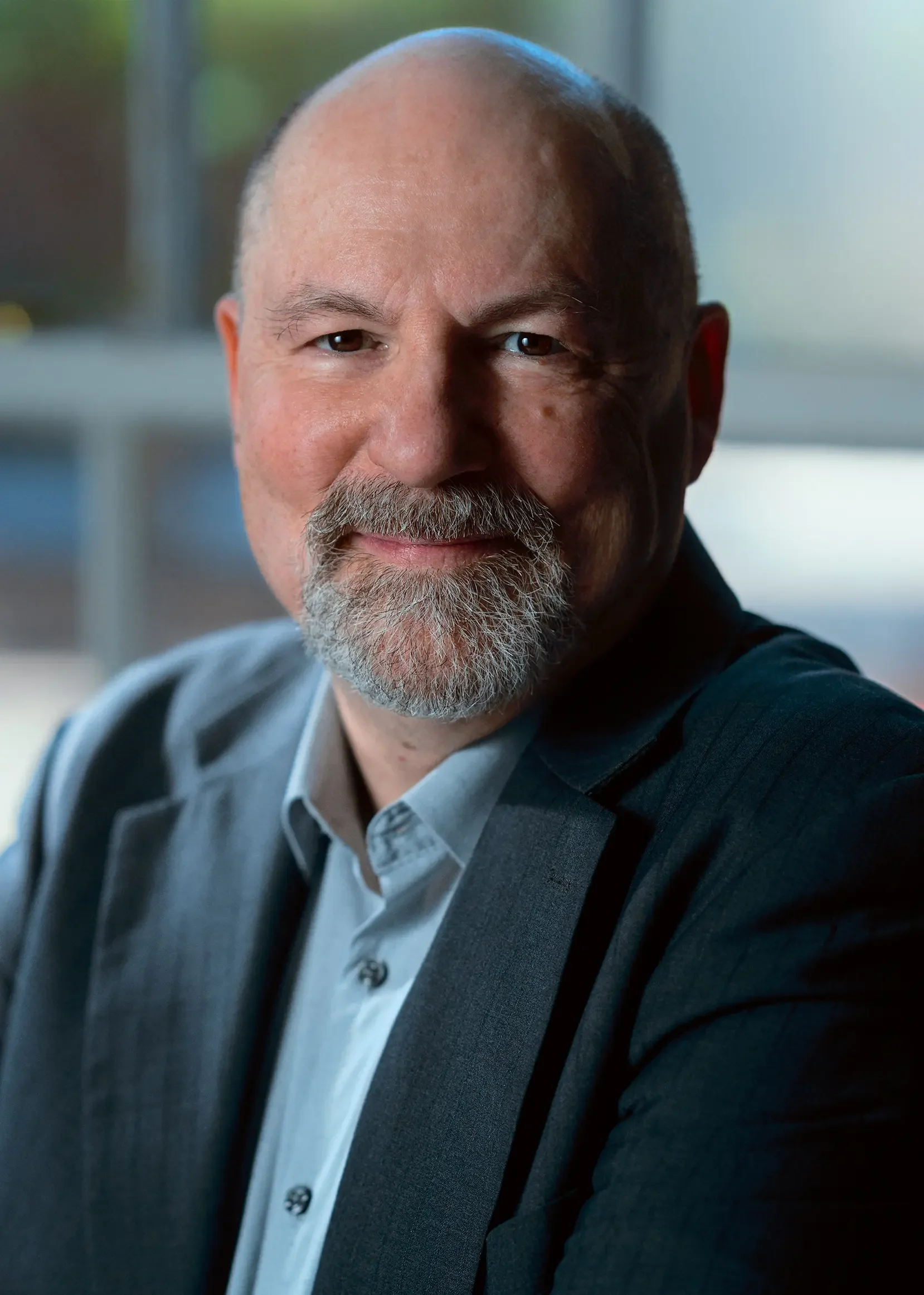 A middle-aged man with a beard and mustache, wearing a light blue shirt and a dark suit, smiling warmly in an office setting | Dr. Robert Meade | Public Speaker & Vision Consultant | Presentations about Eyes and Vision