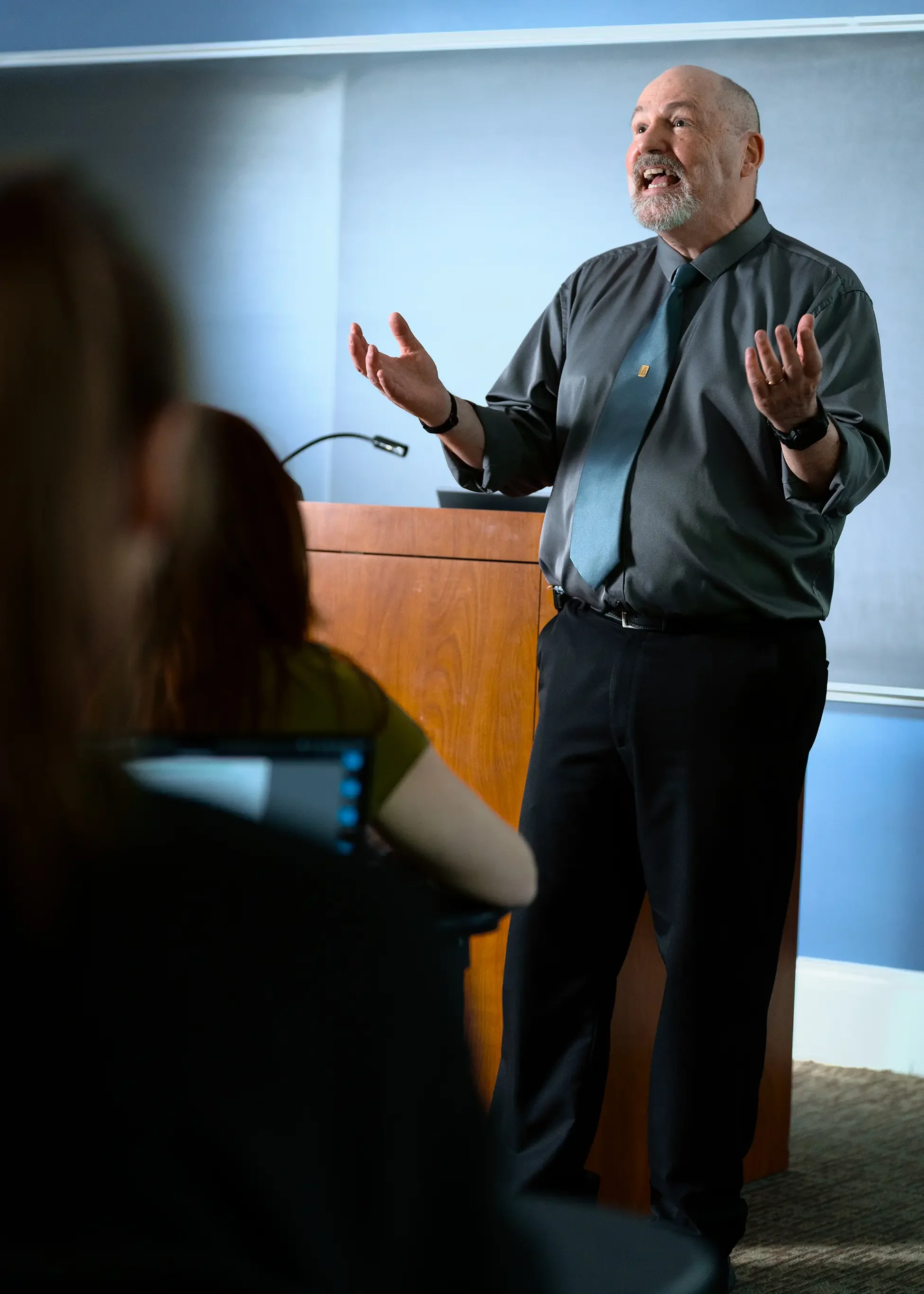 A middle-aged man with a bald head and beard, wearing a dark gray shirt, teal tie, and black pants, is giving a presentation in a classroom with enthusiasm | Dr. Robert Meade | Public Speaker & Vision Consultant | Presentations about Eyes and Vision