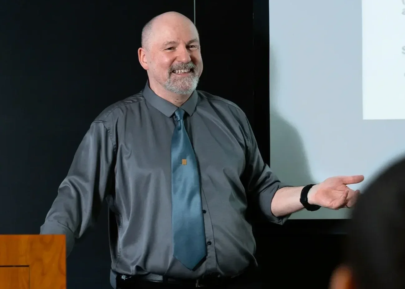 A middle-aged man with a gray beard and bald head, wearing a gray shirt and blue tie, smiling and gesturing with his left hand giving a presentation | Dr. Robert Meade | Public Speaker & Vision Consultant | Presentations about Eyes and Vision