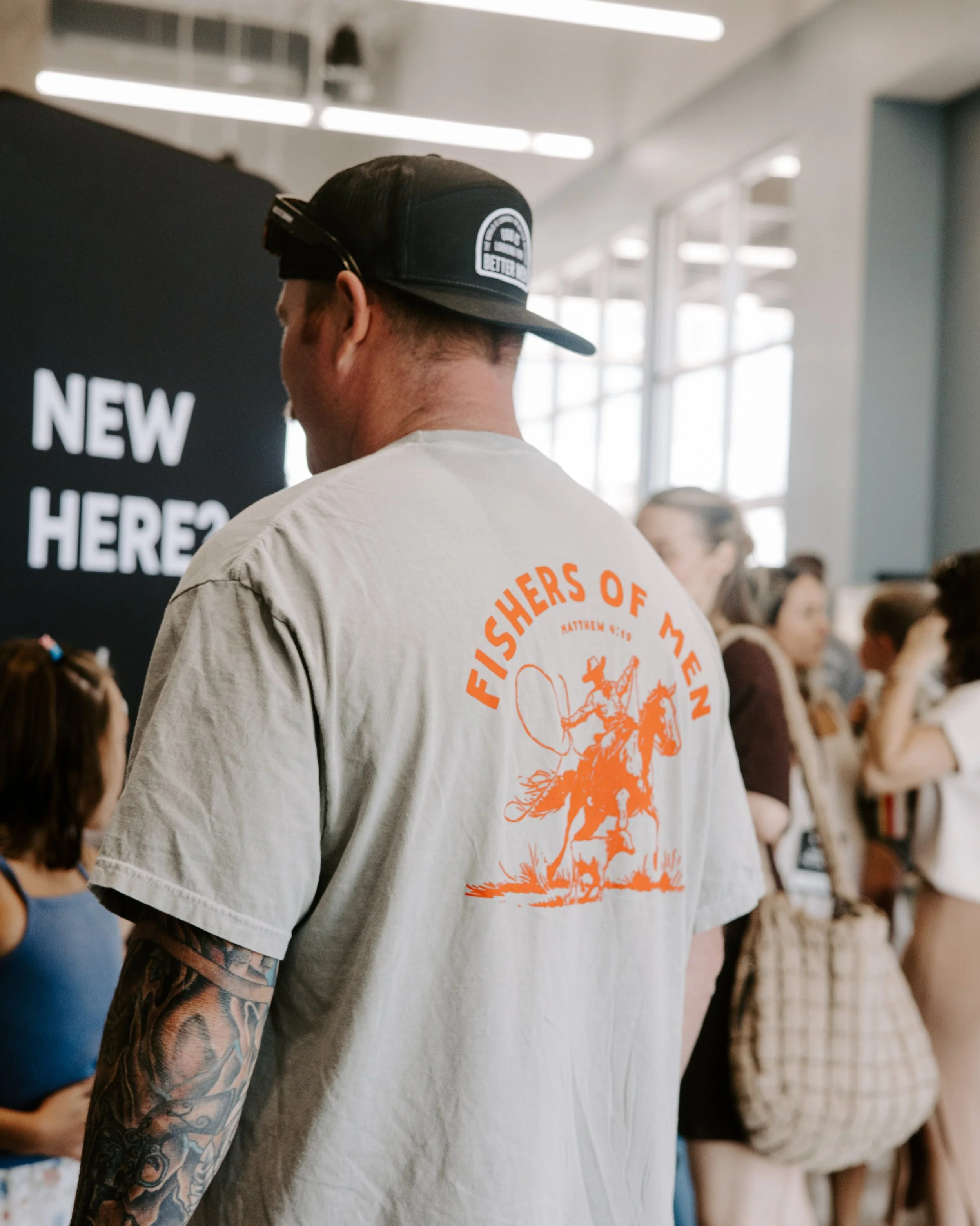 Man wearing a gray T-shirt with an orange graphic of a cowboy on a horse and the text "Fisher of Men". He has a tattoo on his left arm and is wearing a black cap, in a crowded indoor space.