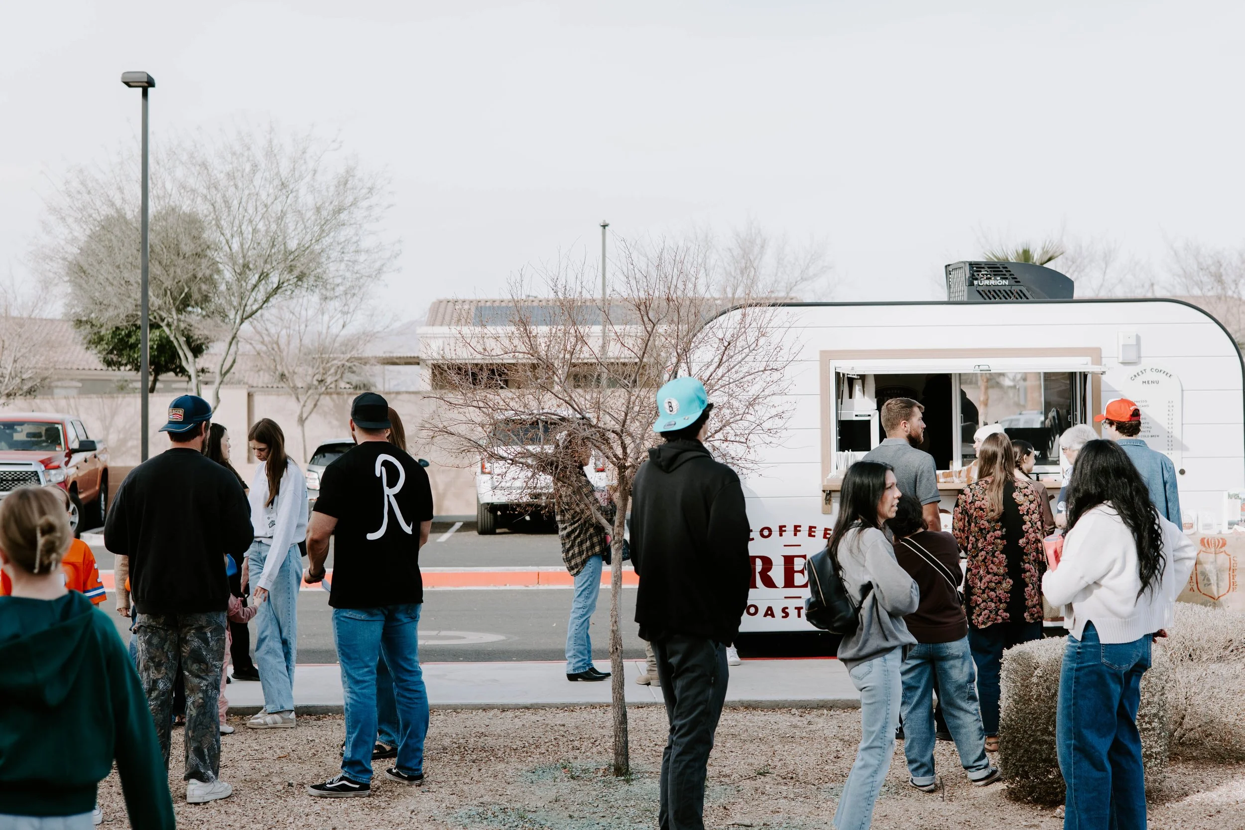 People standing in line at a coffee truck in an outdoor parking lot with leafless trees and parked cars in the background.