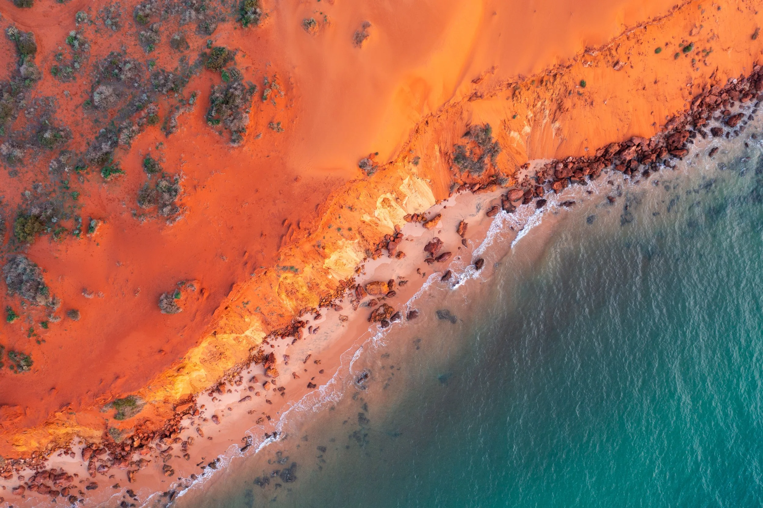 Aerial view of orange-red cliffs meeting a sandy beach, with rocks along the shoreline and ocean water at the bottom edge.