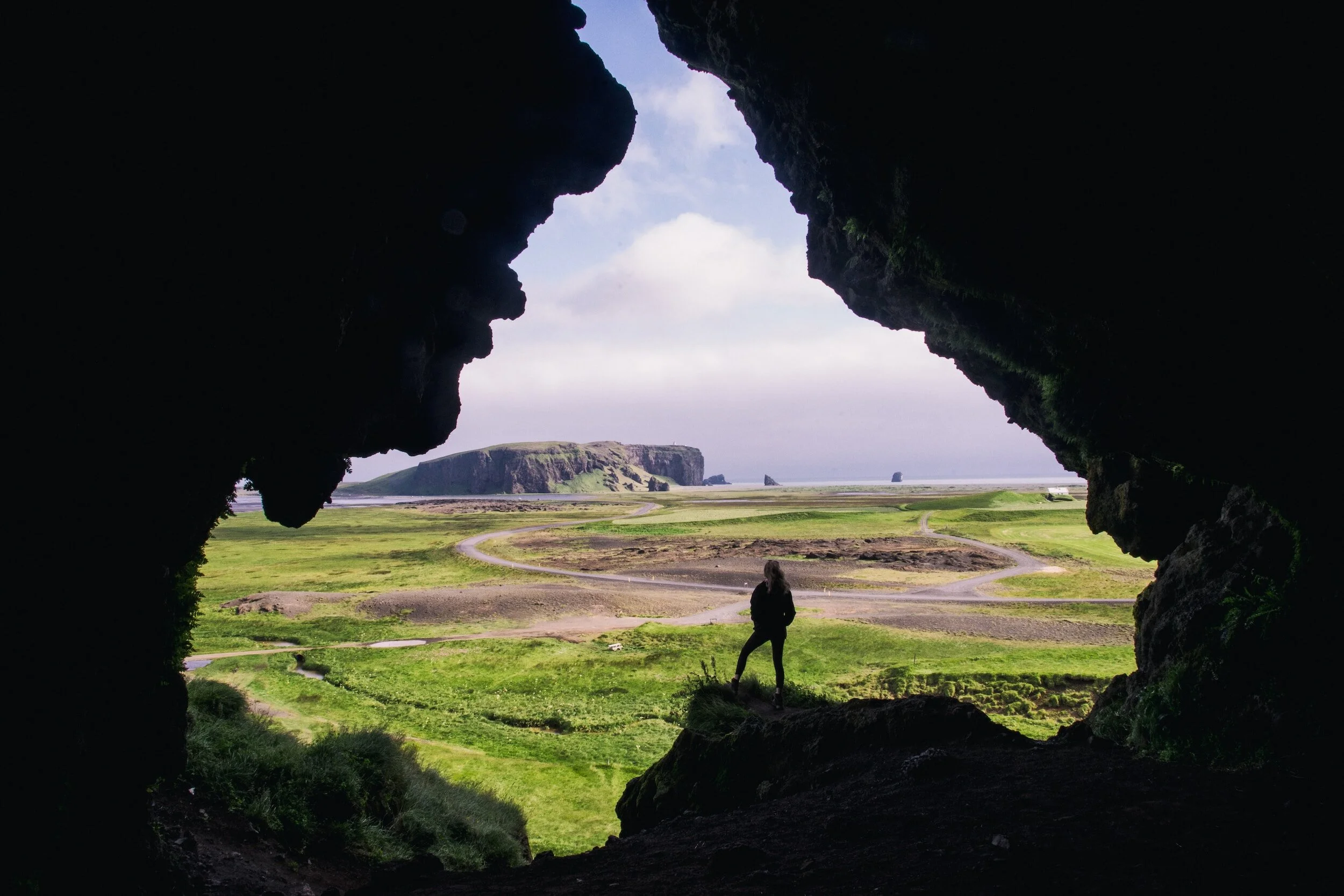 A photographer standing in a cave mouth looking out over green coastal plains