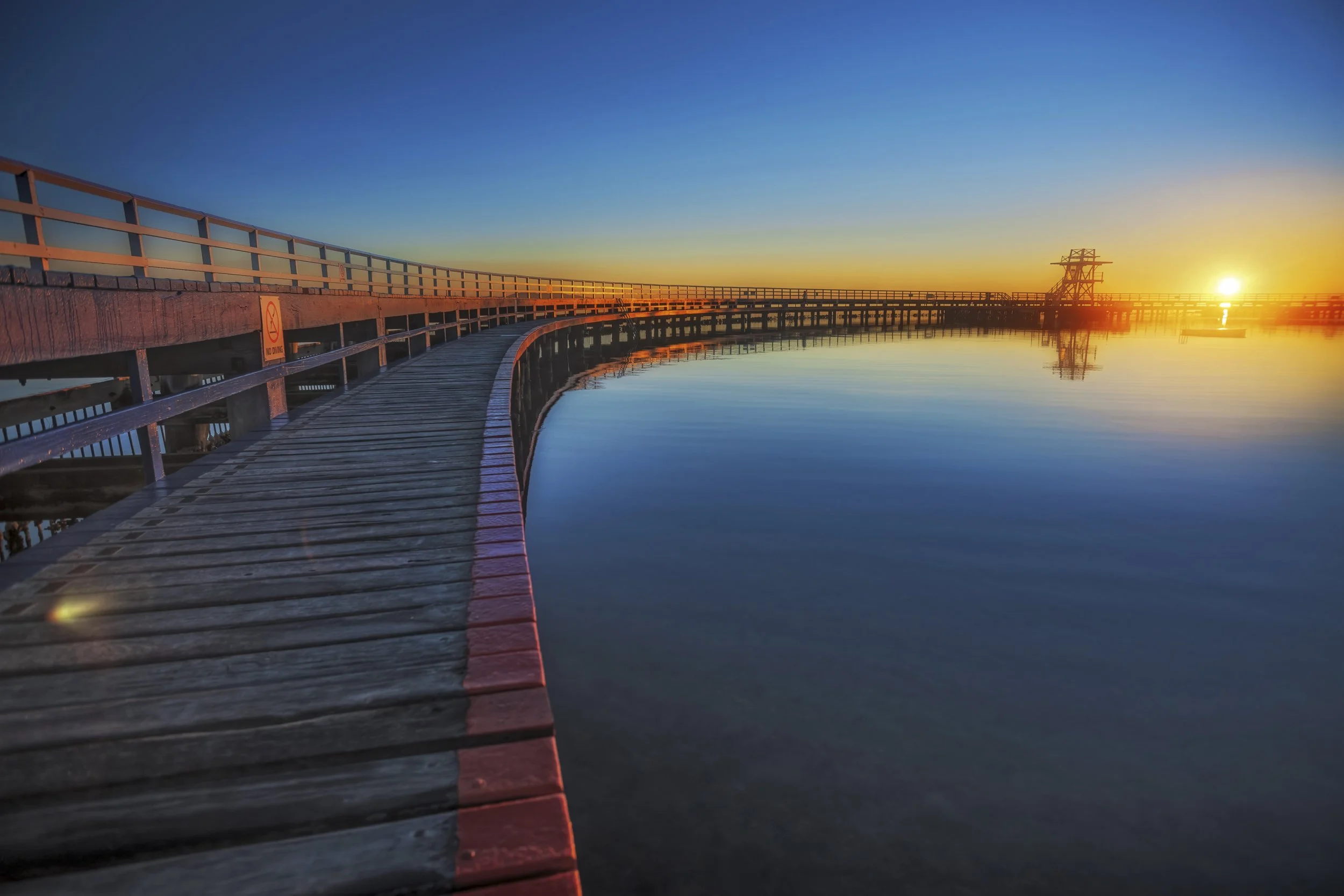A long timber pier stretching into still water with the sun low on the horizon, bathing the scene in golden light.