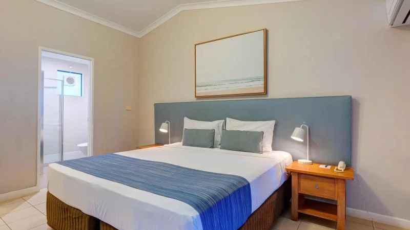 Queen-sized bed with blue bed runner and pillows in a hotel room, wooden nightstand with lamp, wall art depicting a seascape, and a door leading to a bathroom.