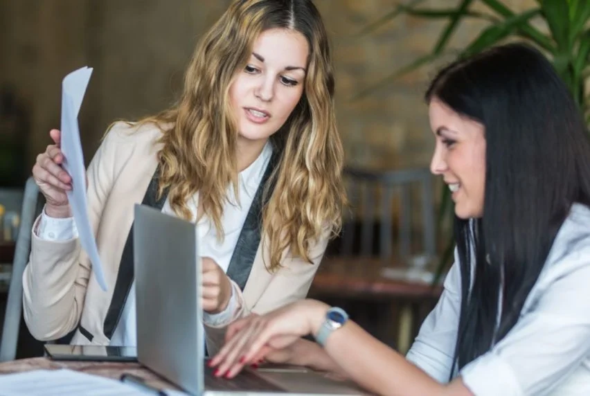 Two young adults sit at right angles to each other. One has long blond hair, the other long black hair. They are looking at the screen of a silver laptop and one is also holding up some pieces of paper that we see from the side.