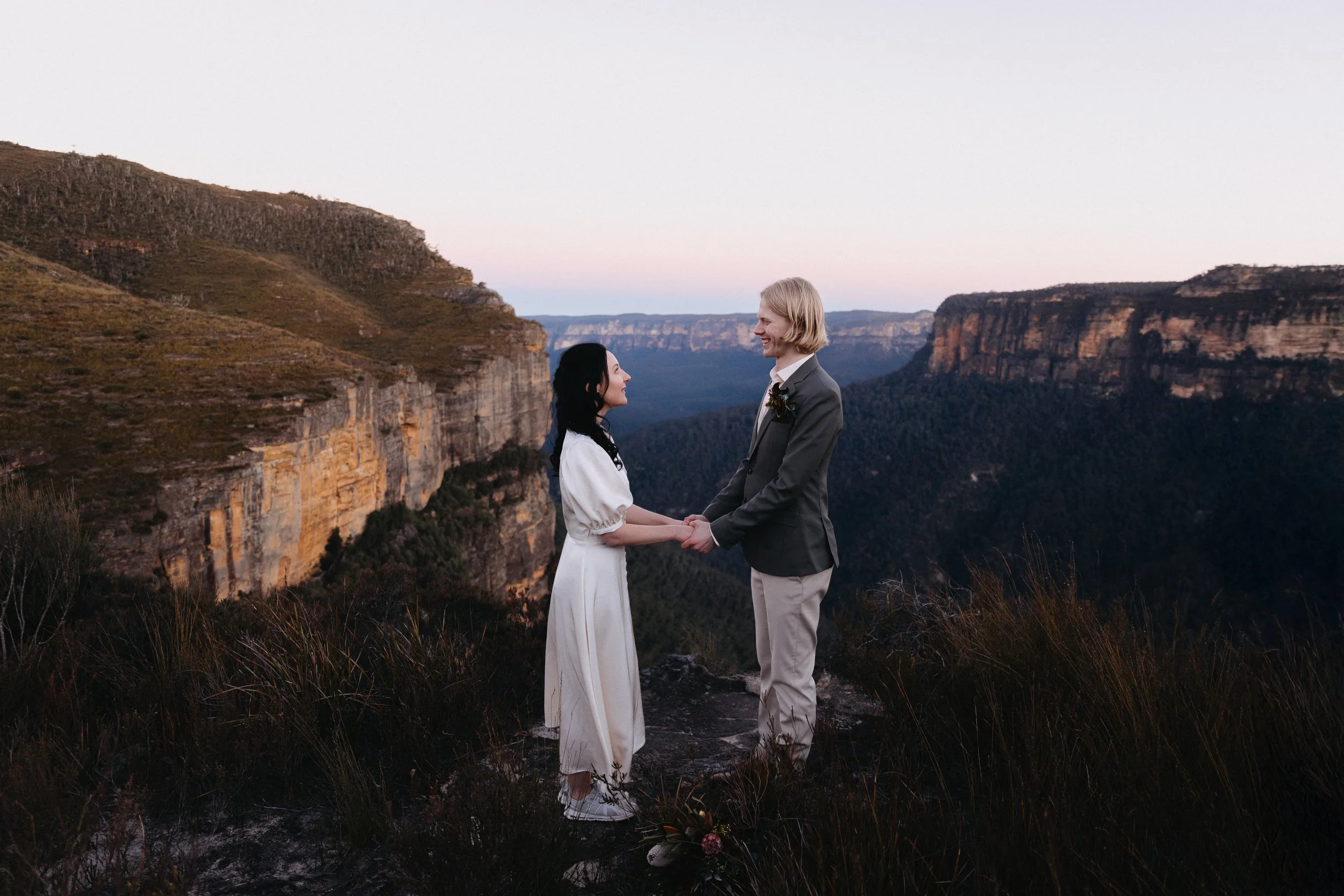 A couple holding hands and smiling at each other on a mountain overlook at sunset with cliffs and valleys in the background.