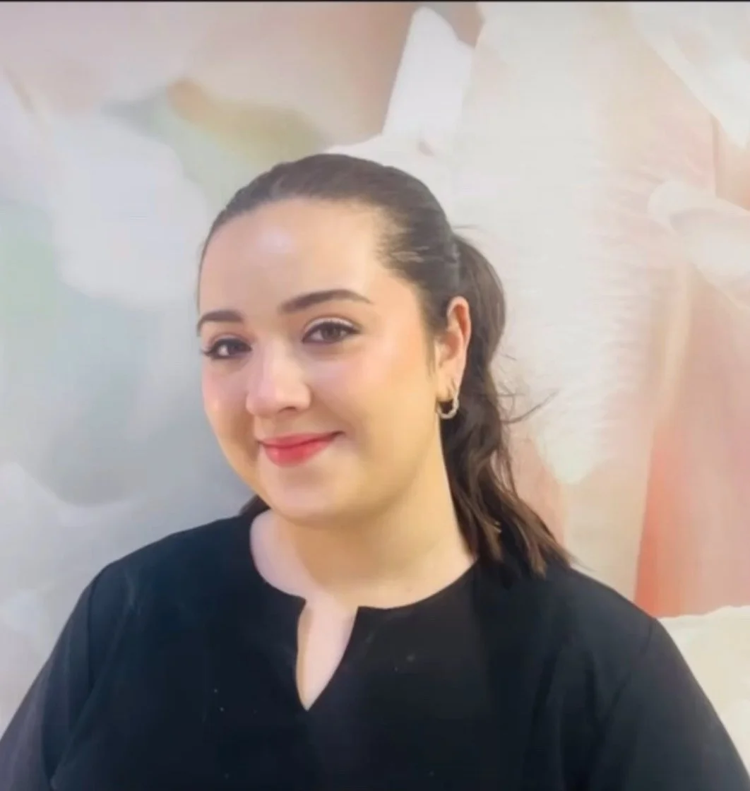 Close-up of a smiling woman with dark hair tied back, wearing a black top and small hoop earrings, against a blurred abstract background.