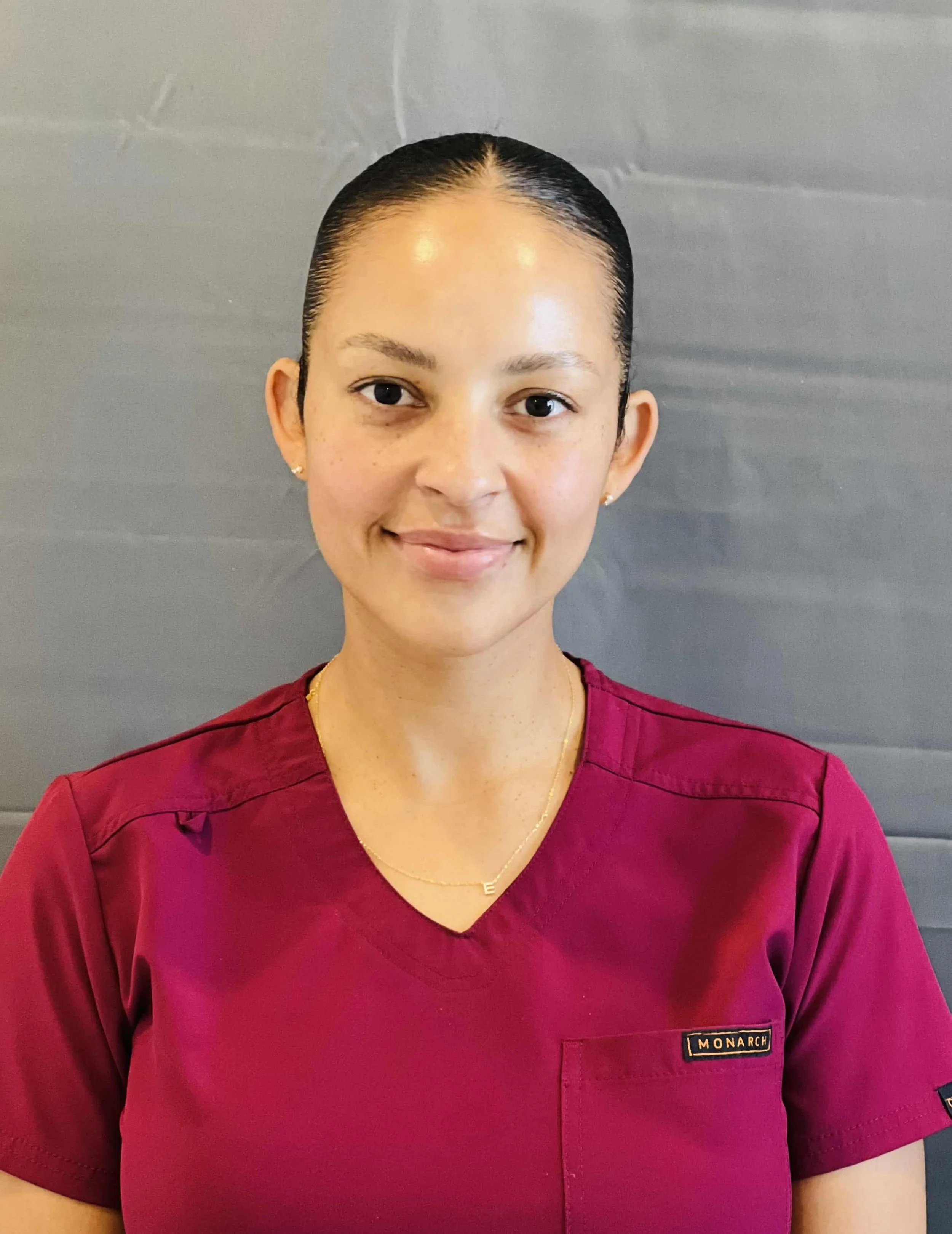 A young woman with dark hair slicked back, wearing a maroon medical uniform with a name tag reading 'MONARCH', standing against a gray background.