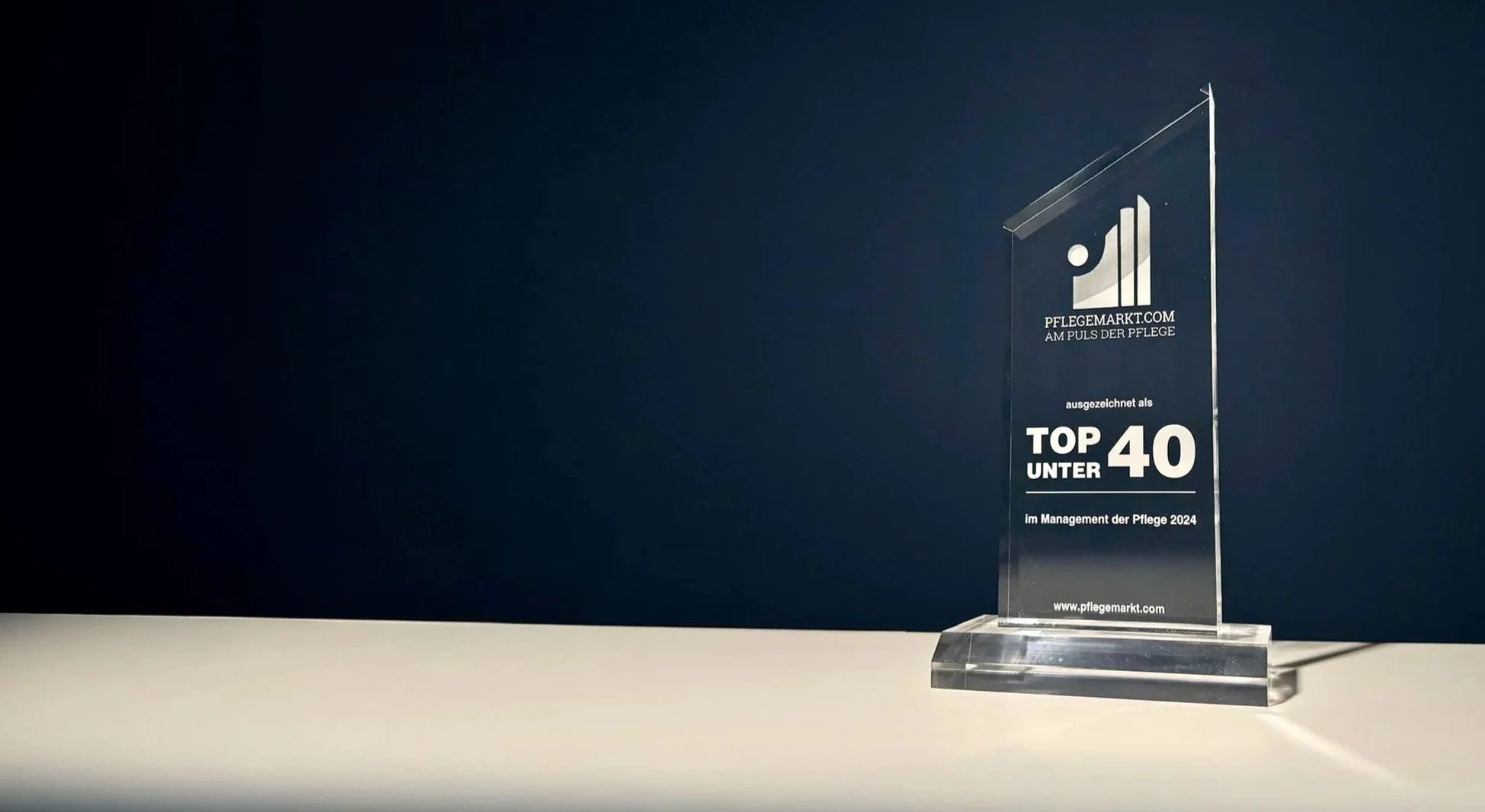 Kristallener Preis für das Pflegeunternehmen, das als Top 40 unter 40 im Management der Pflege 2024 ausgezeichnet wurde, auf weißem Tisch vor dunklem Hintergrund.