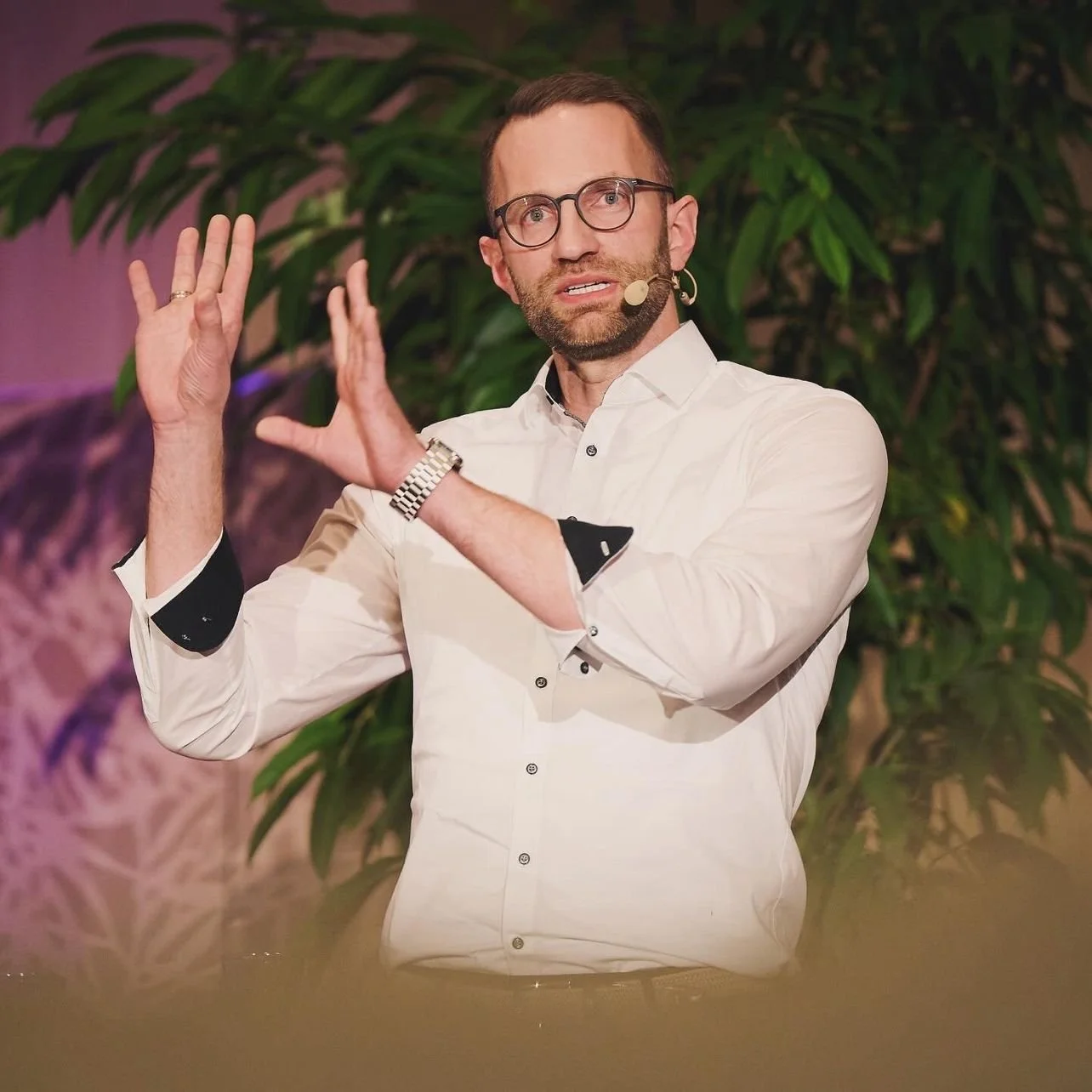 Ein Mann mit Brille und Mikrofon spricht gestikulierend vor einem Hintergrund mit Pflanzen. Er trägt ein weißes Hemd und hat einen Bart.